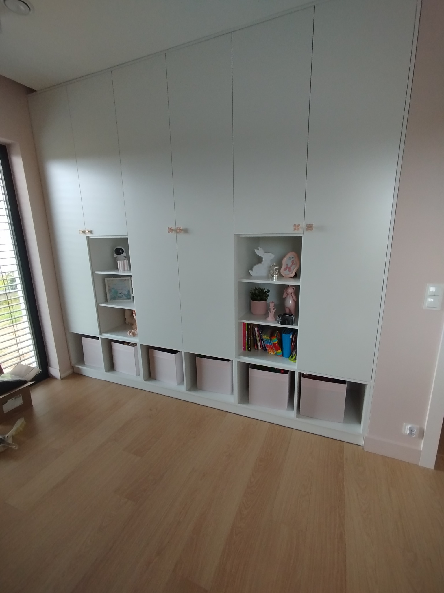 Biała szafa wnękowa z otwartymi półkami i dekoracjami w pokoju dziecięcym. Drzwiczki szafy z delikatnymi, różowymi uchwytami motylkami.