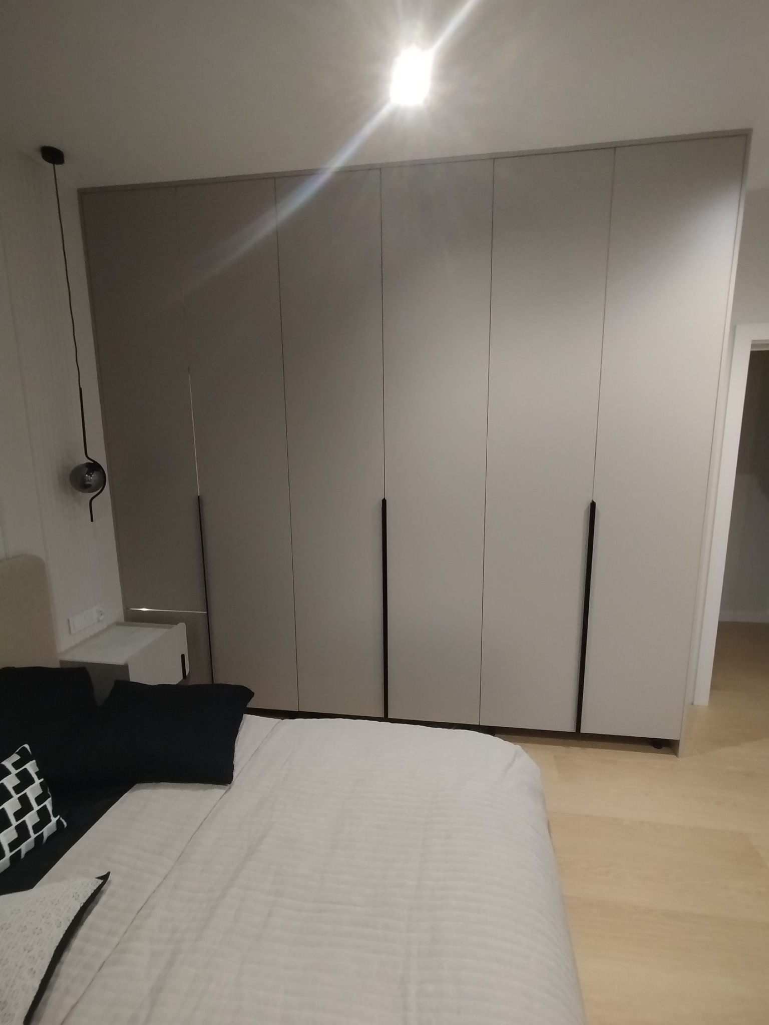 Jasna szafa wnękowa z długimi, czarnymi uchwytami, umieszczona w sypialni z widocznym łóżkiem i lampką nocną. Minimalistyczny design.