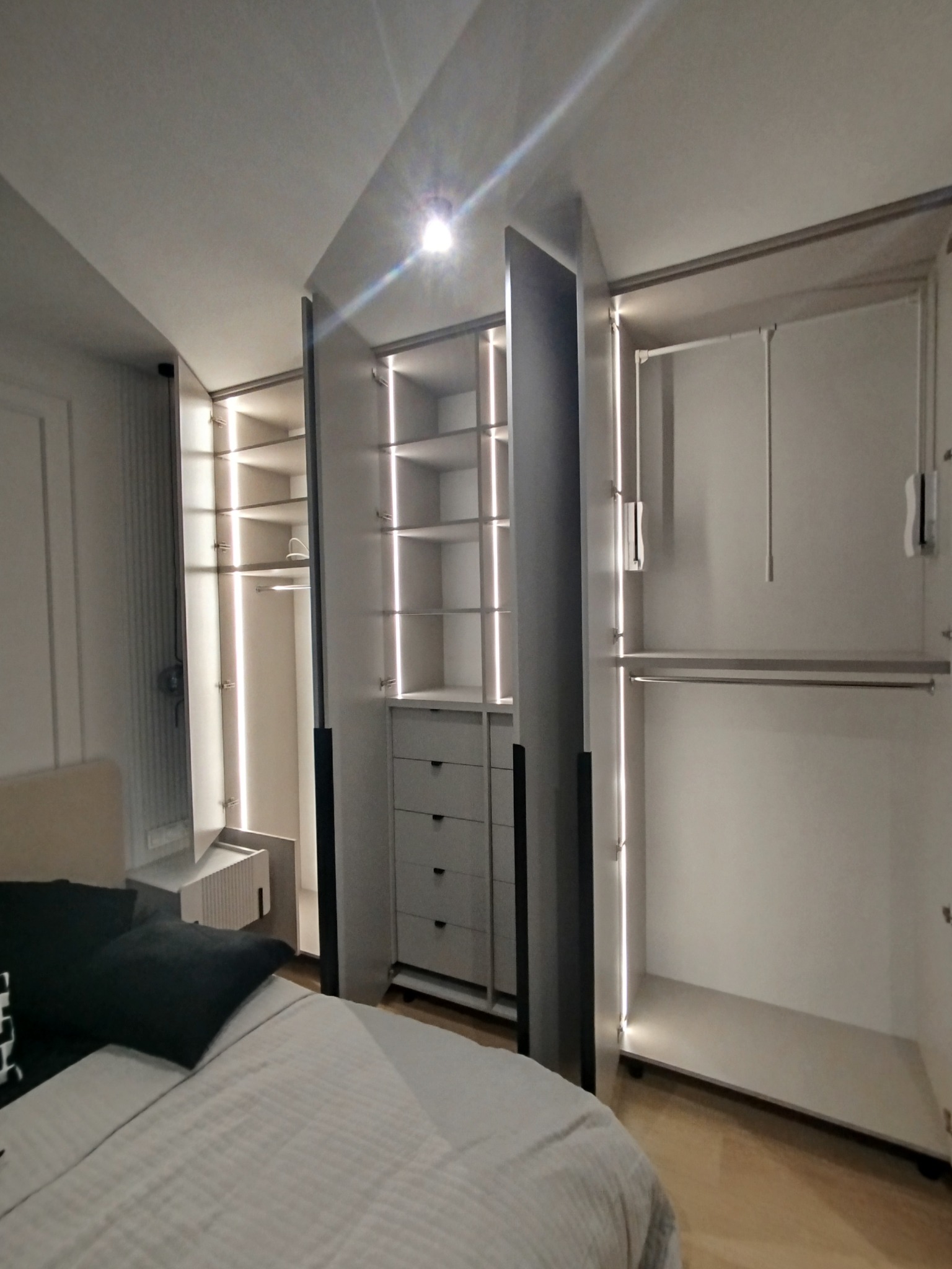 Nowoczesna szafa wnękowa z oświetleniem LED, widoczna z perspektywy łóżka. Jasne wnętrze szafy z półkami i szufladami. Minimalistyczny design w sypialni.