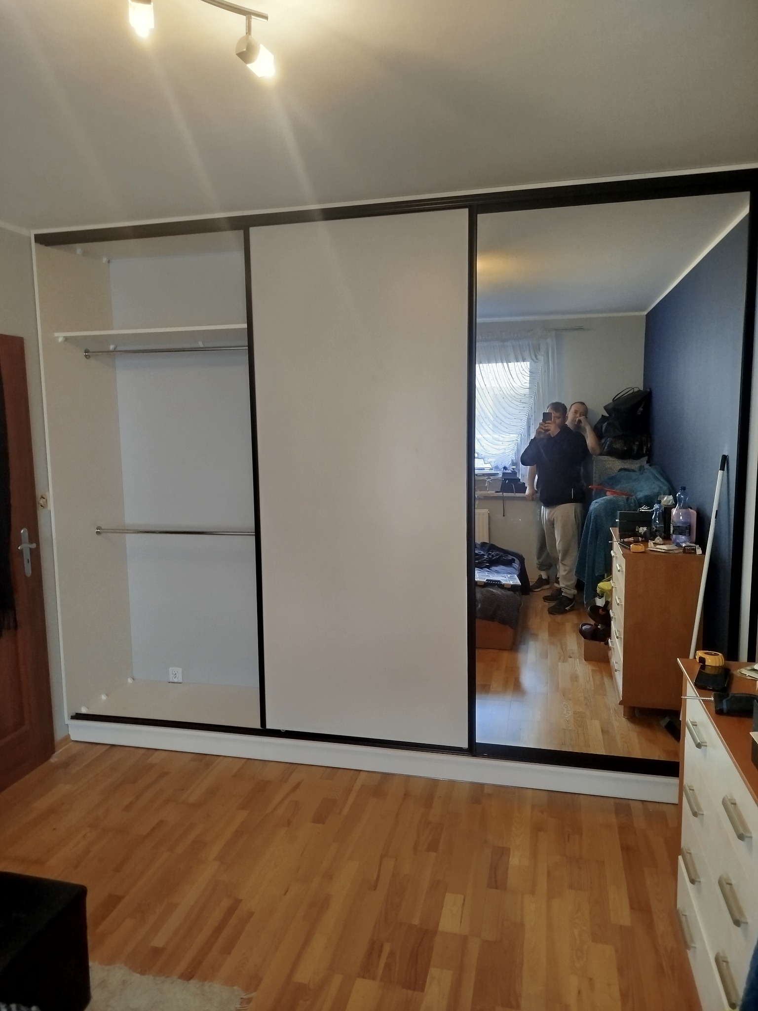 Szafa wnękowa z przesuwnymi drzwiami, z lustrem odbijającym wnętrze pokoju z dwoma osobami. Widoczne półki i drążki w szafie, parkiet na podłodze.