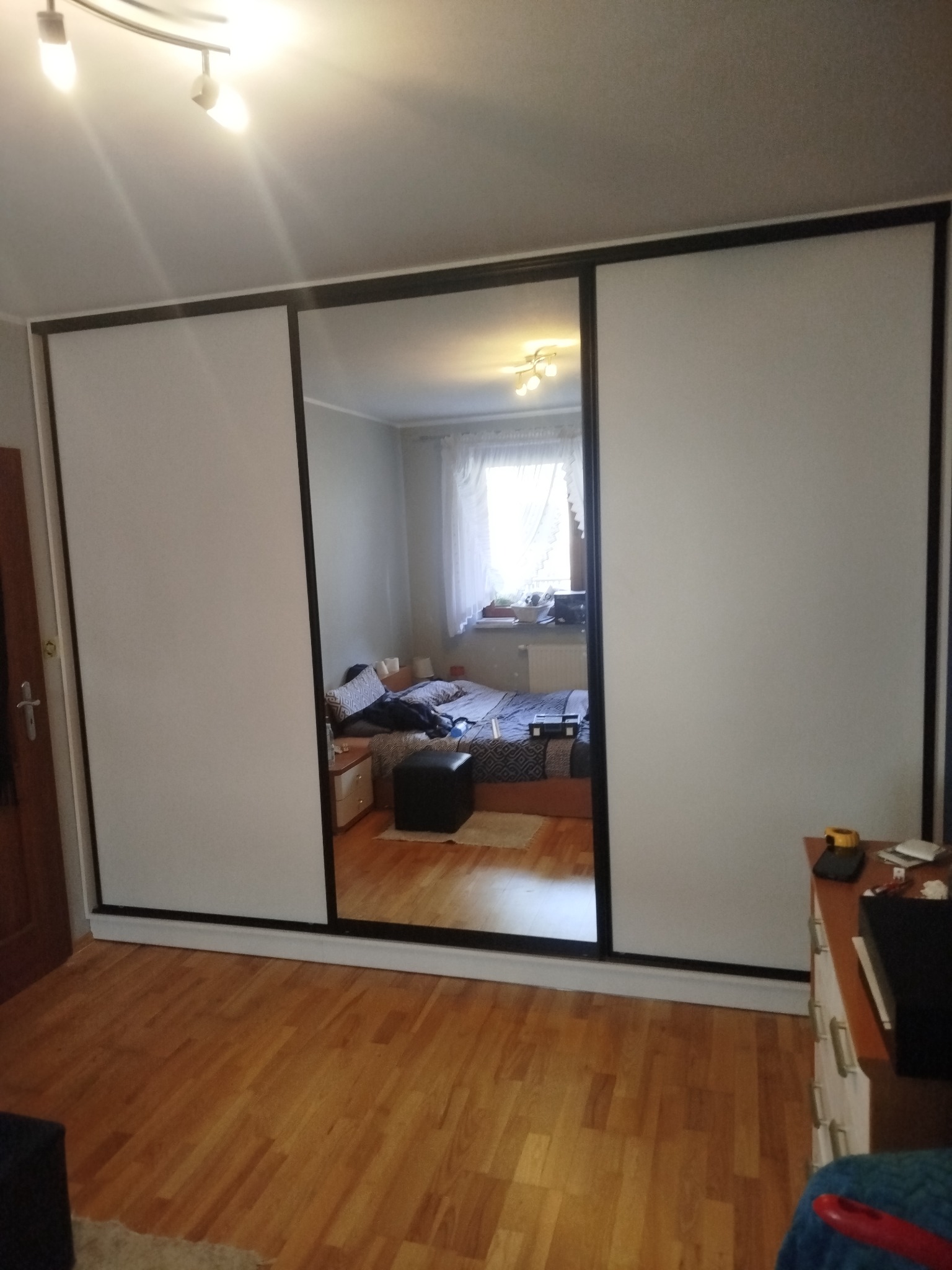 Szafa przesuwna z lustrzanymi drzwiami w pokoju, odbicie wnętrza sypialni. Minimalistyczny design, jasne fronty, czarne obramowania. Parkiet na podłodze.