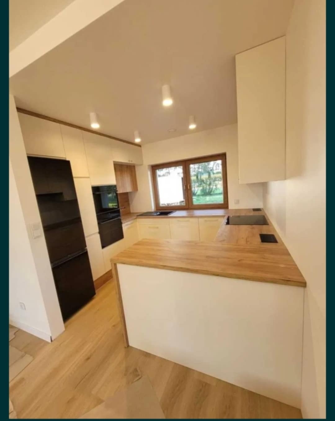 Jasna kuchnia z drewnianym blatem i oknem. Widoczny czarny sprzęt AGD, oświetlenie punktowe i drewniana podłoga. Nowoczesny design.