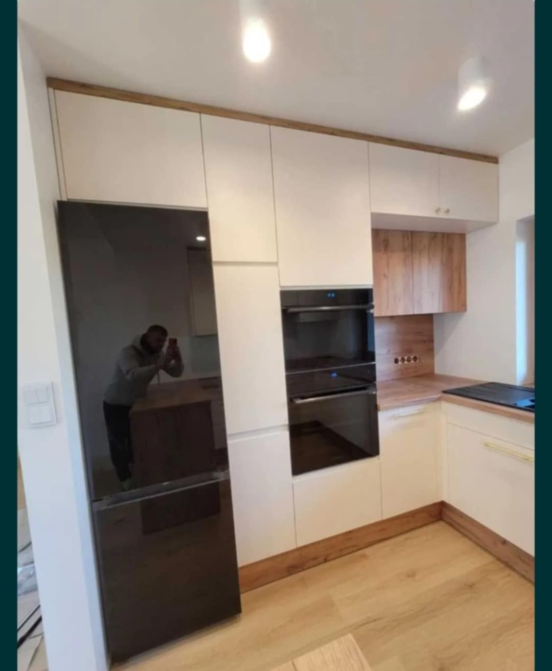 Jasna kuchnia z drewnianymi akcentami: zabudowa z lodówką w czarnym połysku i piekarnikiem. Na blacie płyta grzewcza. W odbiciu widać fotografa.