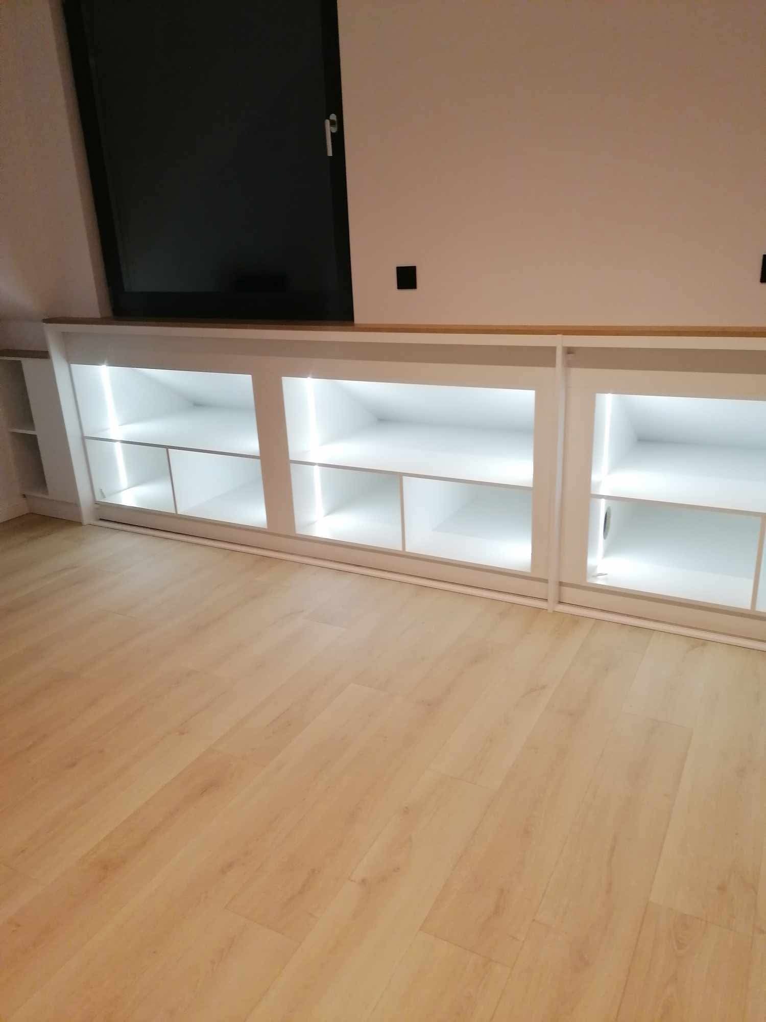 Biała szafa wnękowa pod skosem dachu z oświetleniem LED, drewniany blat, jasna podłoga. Minimalistyczny design.