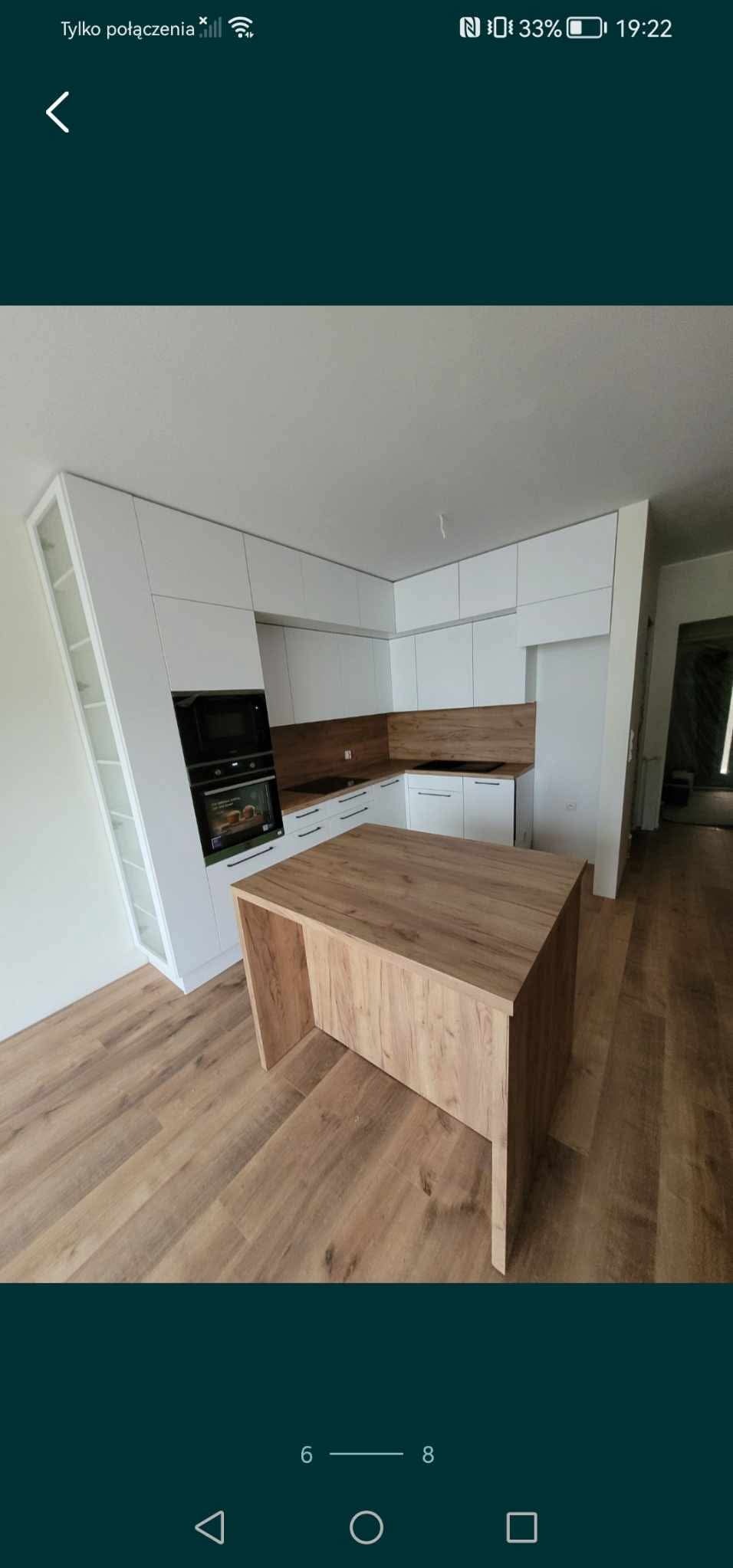 Nowoczesna kuchnia z białą zabudową i wyspą z blatem drewnopodobnym. Widoczne sprzęty AGD w zabudowie, minimalistyczny design.