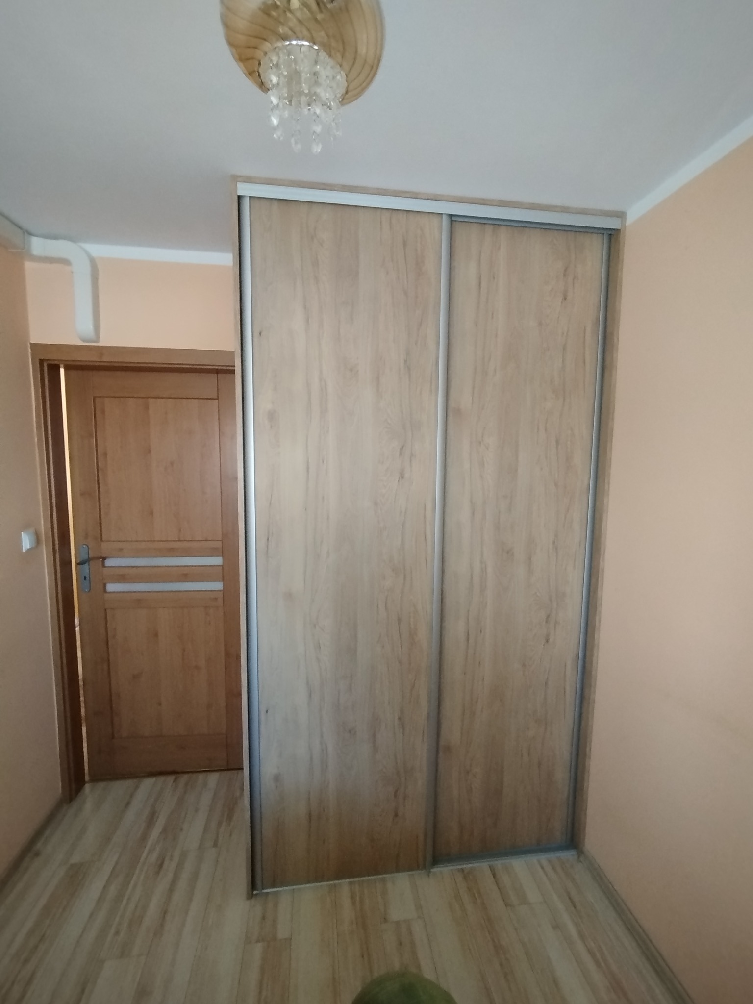 Jasna szafa wnękowa z przesuwnymi drzwiami o drewnopodobnym wzorze, dopasowana do wnętrza pokoju z drewnianą podłogą i drzwiami. Widoczny fragment ozdobnego żyrandola.