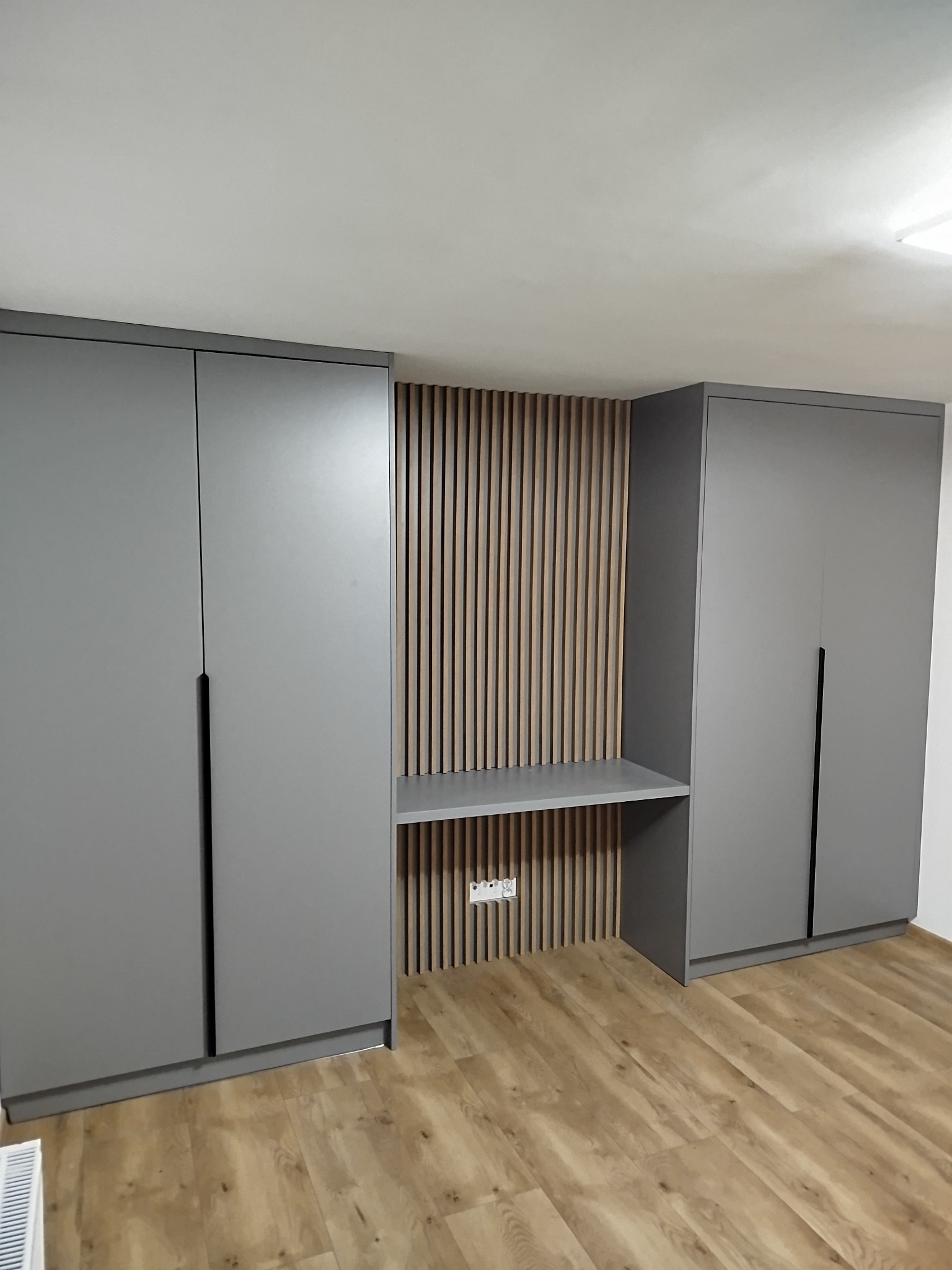Szara szafa wnękowa z biurkiem i drewnianymi lamelami na ścianie, widok na podłogę z paneli. Minimalistyczny design, nowoczesne wykończenie.