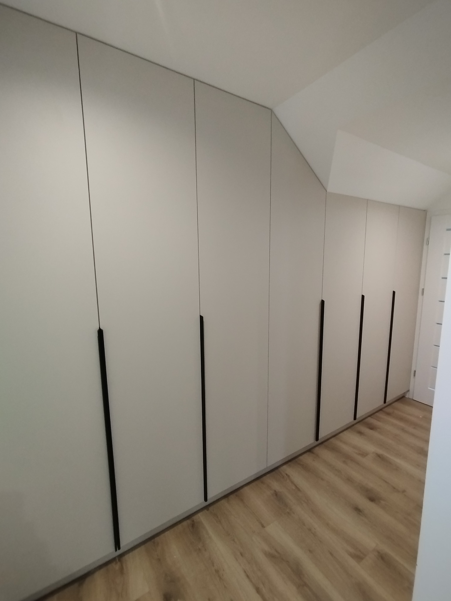 Długa, zabudowana szafa z jasnymi frontami i czarnymi, pionowymi uchwytami, dopasowana do skosu sufitu, z widoczną podłogą w odcieniach drewna.