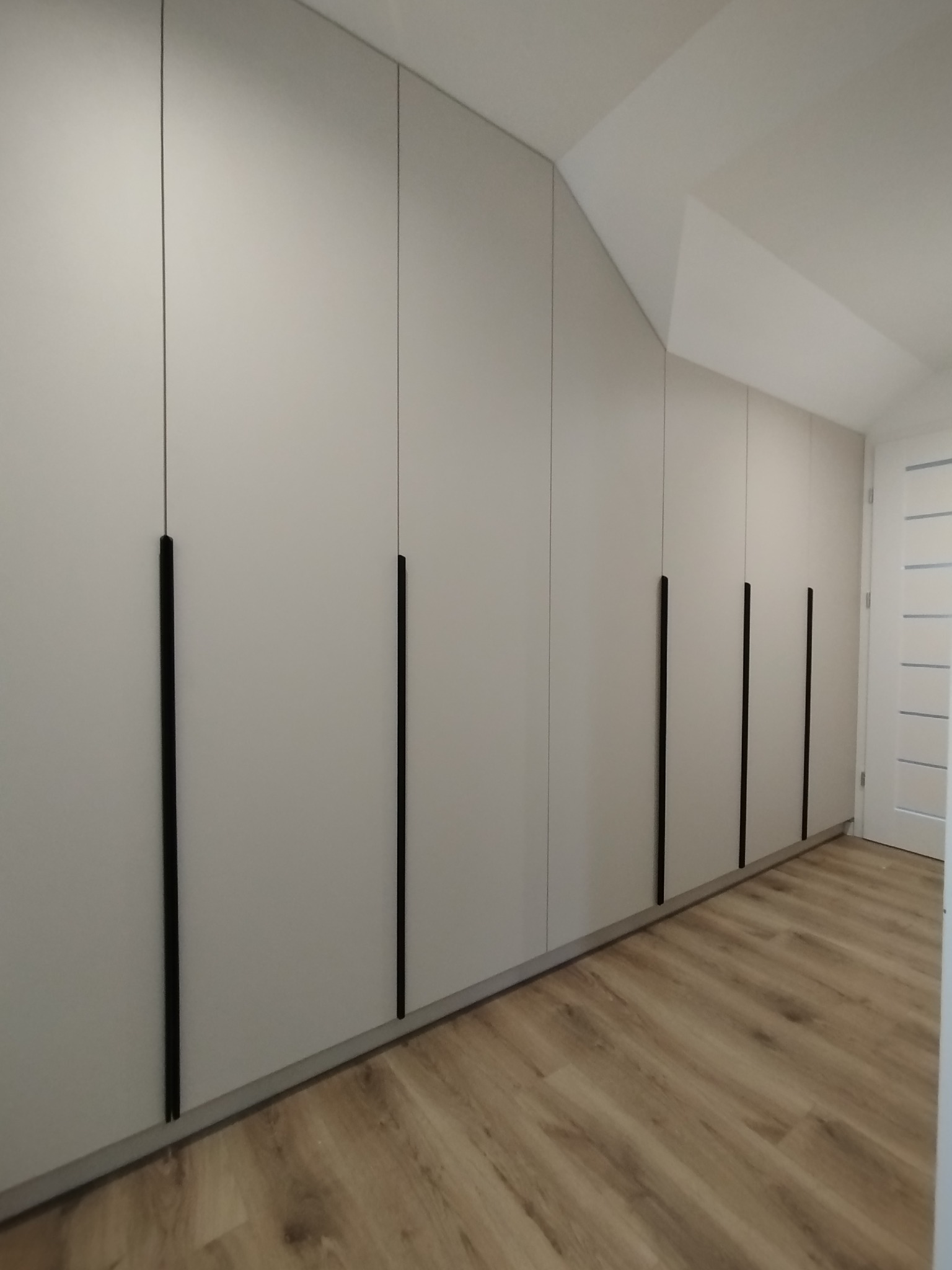 Biała szafa wnękowa z długimi, czarnymi uchwytami, dopasowana do skosu dachu w Twardogórze. Jasne panele na podłodze i białe drzwi w tle.