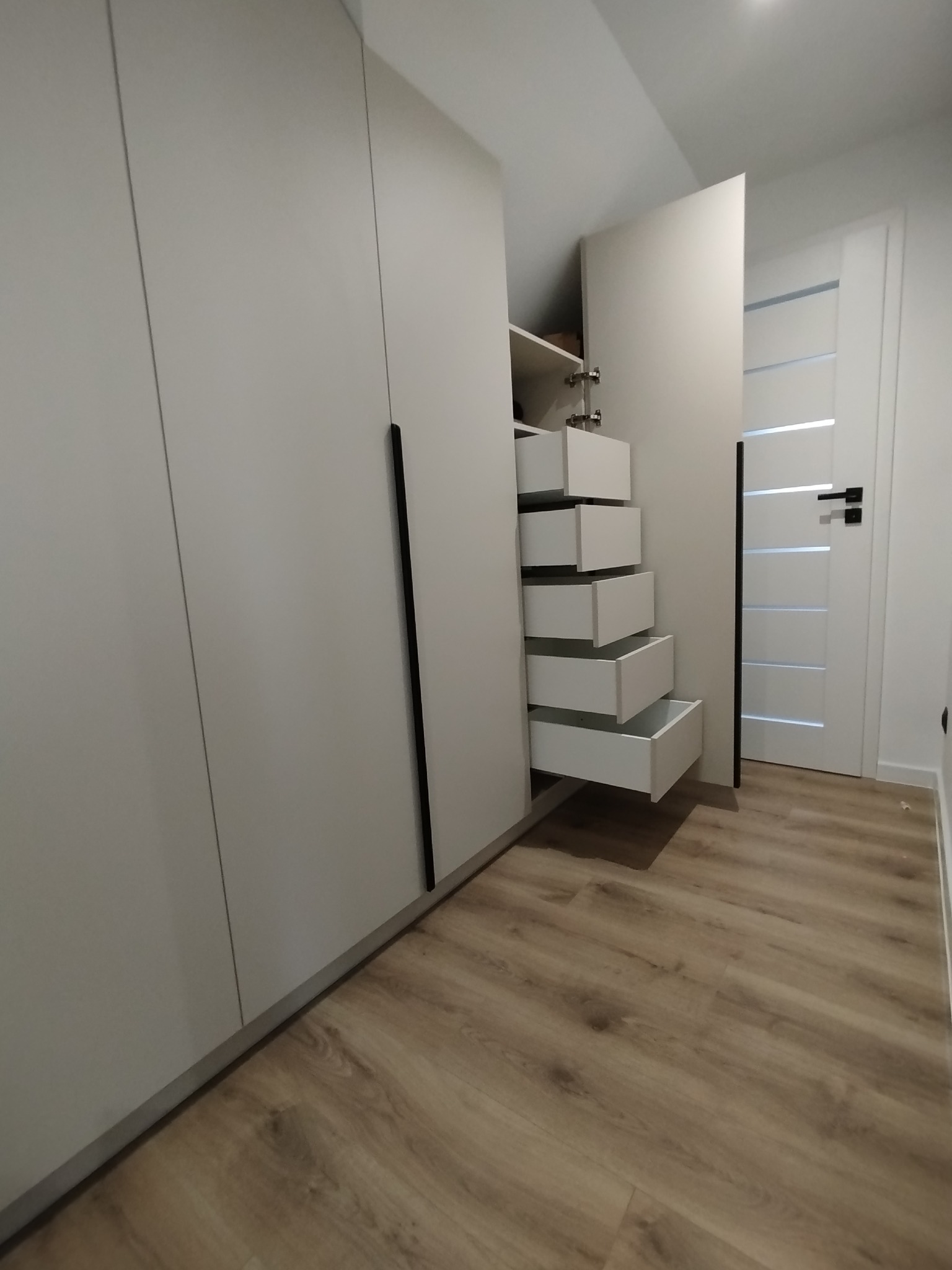 Biała szafa wnękowa z czarnymi uchwytami, otwarte szuflady wewnątrz. Widoczna podłoga z paneli i białe drzwi z przeszkleniem.