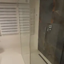 Nowoczesna kabina prysznicowa typu walk-in z szklaną ścianką, deszczownicą i odpływem liniowym w posadzce, na tle ściany wykończonej płytami imitującymi kamień.