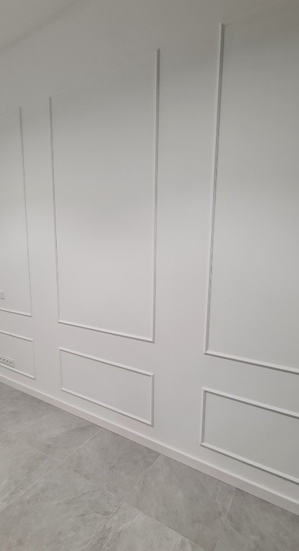 Ściana z dekoracyjnymi listwami w kolorze białym w mieszkaniu w Gorzowie Wielkopolskim, widoczne szare płytki podłogowe.