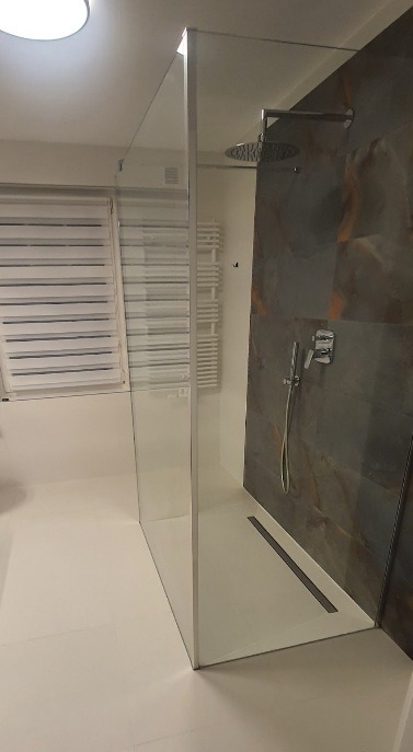 Nowoczesna kabina prysznicowa typu walk-in z szklaną ścianką, deszczownicą i odpływem liniowym w posadzce, na tle ściany wykończonej płytami imitującymi kamień.