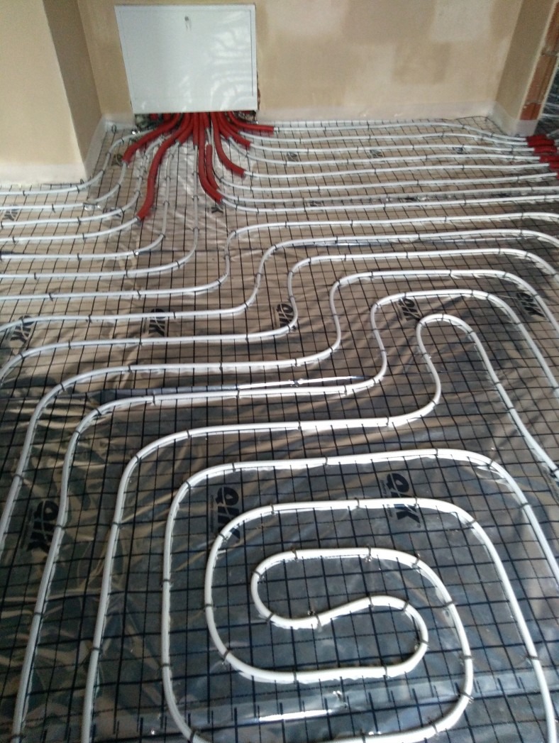 Białe rury ogrzewania podłogowego ułożone na czarnej siatce, z czerwonymi rurami przy ścianie. Widoczny fragment ściany i biała skrzynka instalacyjna.