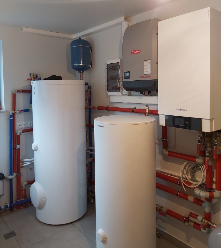 Zestaw urządzeń grzewczych w kotłowni: dwa białe zbiorniki, niebieski zasobnik, pompa ciepła Fronius i piec Viessmann. Instalacja z rur czerwonych i niebieskich.