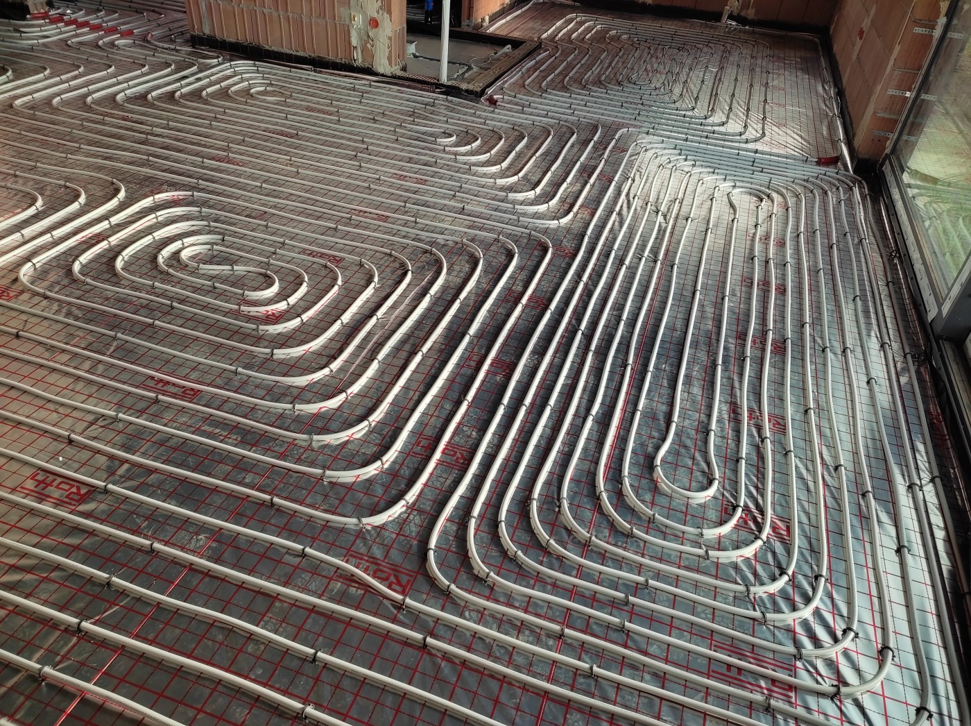 Białe rury ogrzewania podłogowego ułożone na czerwonej siatce, widoczne fragmenty ścian z cegły. Układ spiralny i równoległy rur. Etap instalacji przed wylewką.