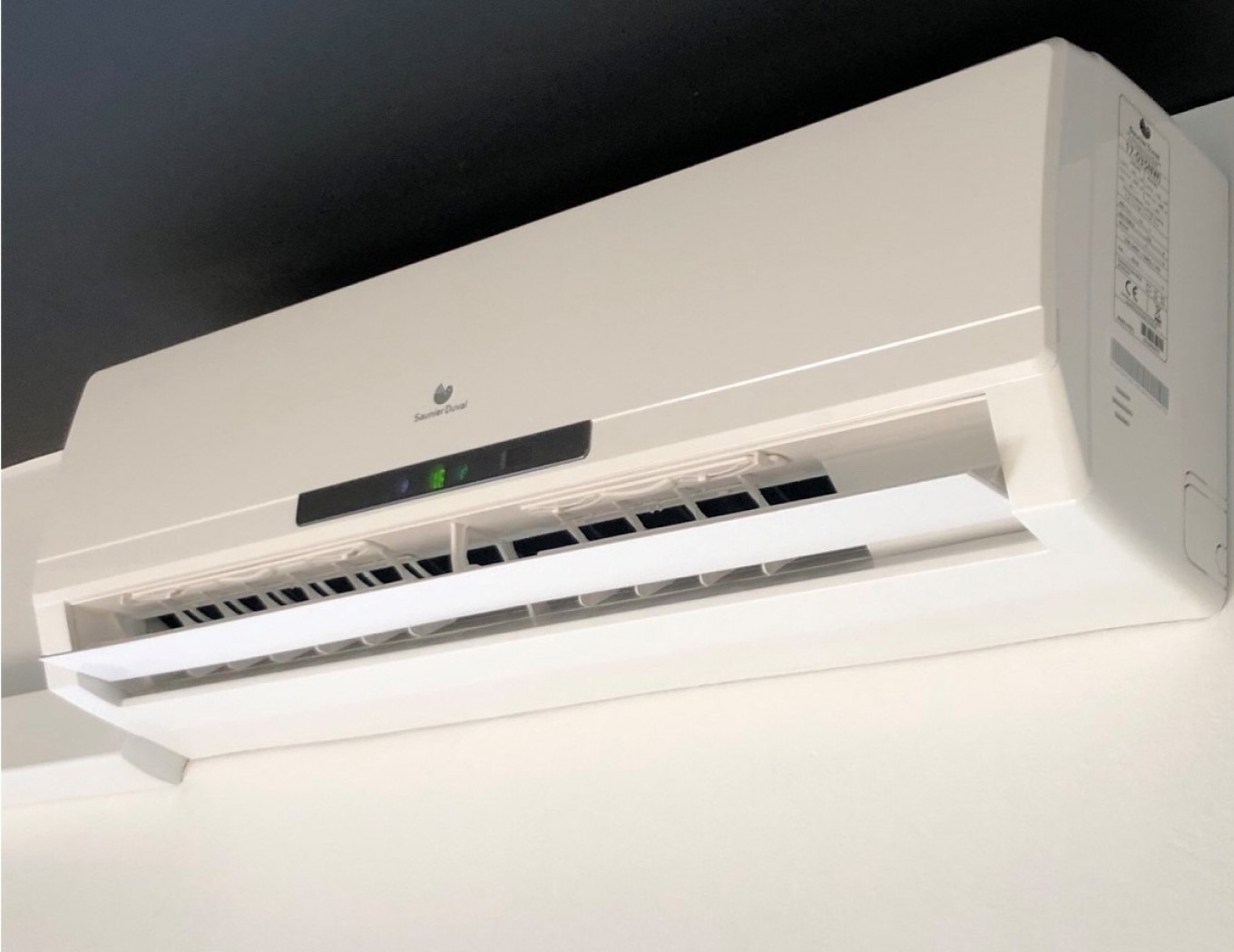 Biała, naścienna pompa ciepła marki Saunier Duval zamontowana pod ciemnym sufitem. Widoczne detale urządzenia i etykieta z parametrami.