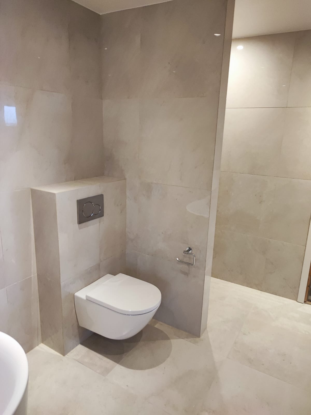 Beżowa łazienka z podwieszaną toaletą i ukrytą spłuczką. Minimalistyczny design, płytki imitujące kamień. Widoczne detale wykończenia, czysta i nowoczesna aranżacja.