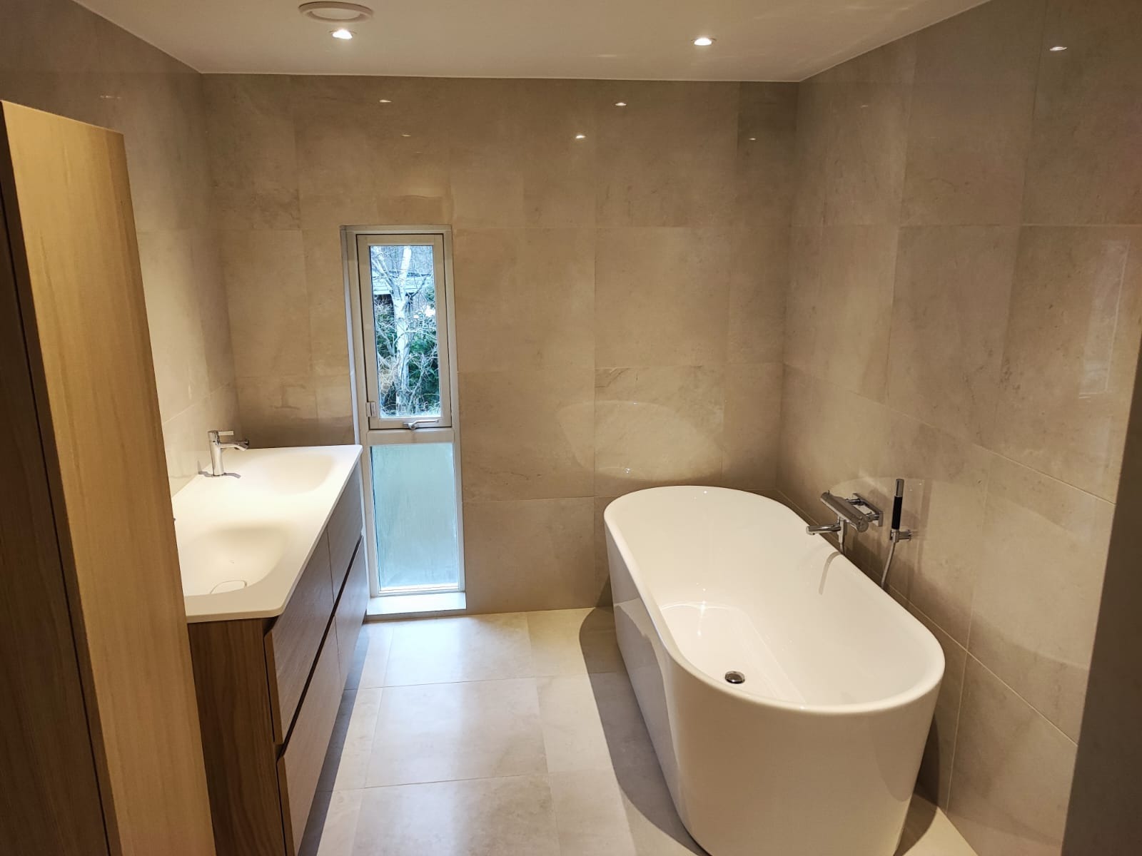 Nowoczesna łazienka w beżowych płytkach z wolnostojącą wanną, umywalką z szafką z drewna i wąskim oknem z widokiem na zieleń na zewnątrz.