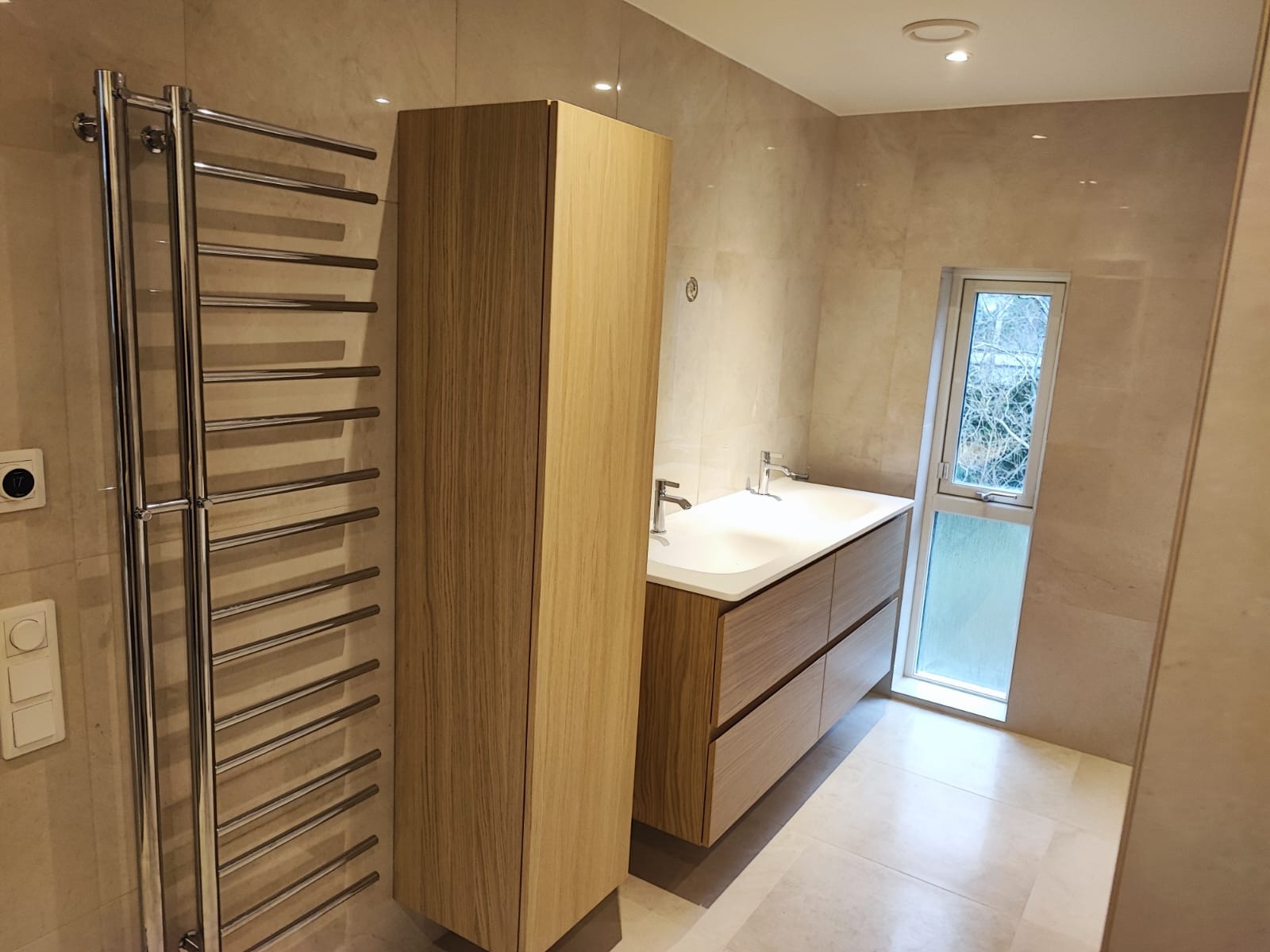 Wykończona łazienka w beżowych płytkach z drewnianymi szafkami, grzejnikiem drabinkowym i oknem. Widok na umywalkę z dwiema bateriami i wysoką szafkę.