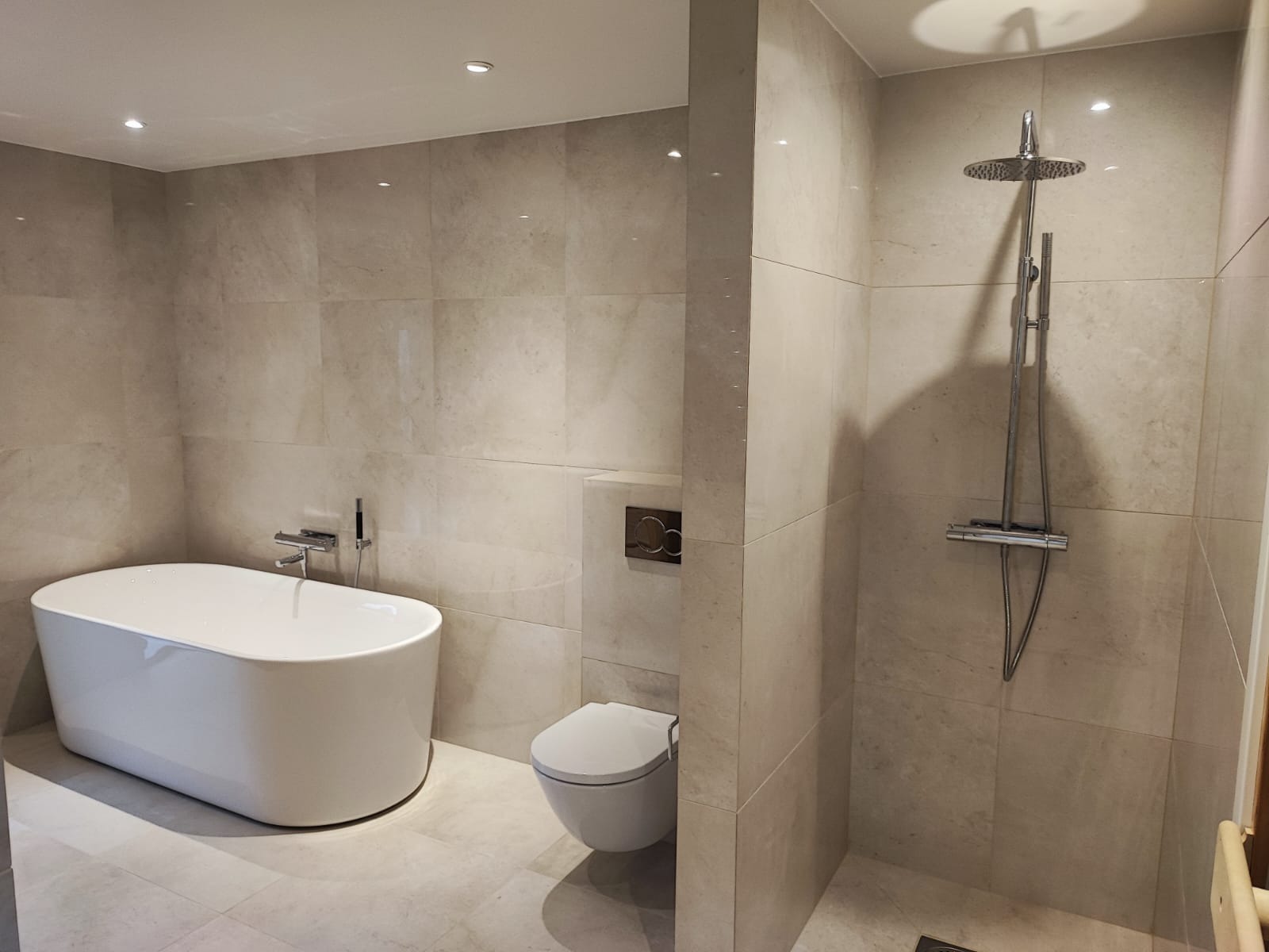 Nowoczesna łazienka z wanną wolnostojącą, toaletą podwieszaną i kabiną prysznicową typu walk-in, wykończona beżowymi płytkami wielkoformatowymi. Minimalistyczny design.