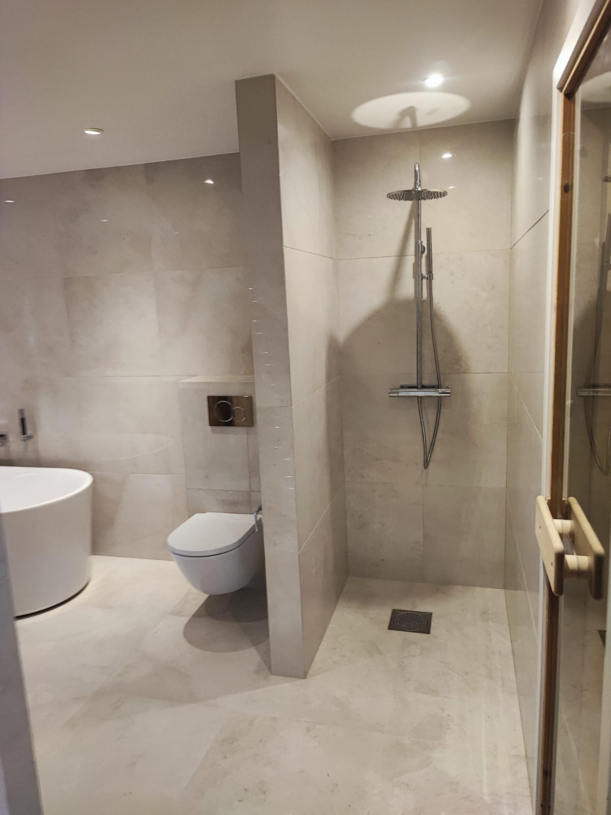 Jasna łazienka z kabiną prysznicową typu walk-in, wiszącą toaletą i wolnostojącą wanną. Płytki w odcieniach beżu, minimalistyczny design, punktowe oświetlenie.