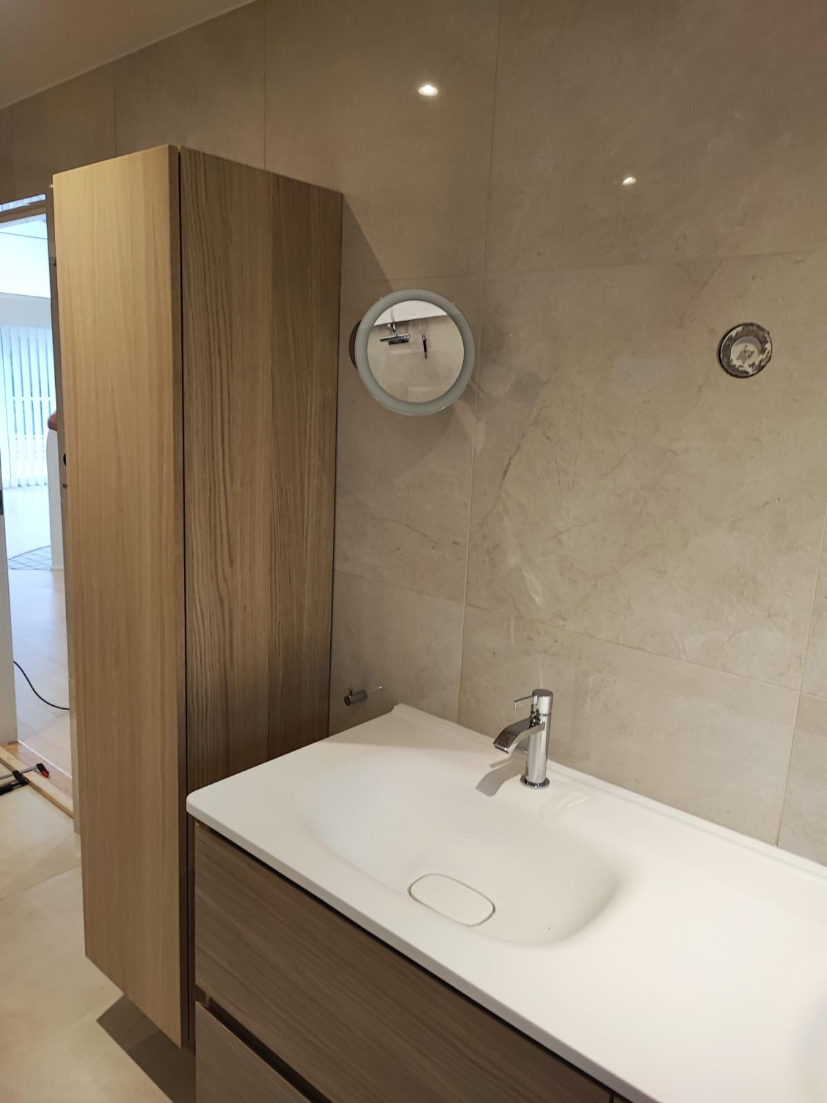 Nowoczesna łazienka w Kaliskach z drewnianą szafką pod umywalką, okrągłym lustrem z podświetleniem i beżowymi płytkami. Minimalistyczny design.