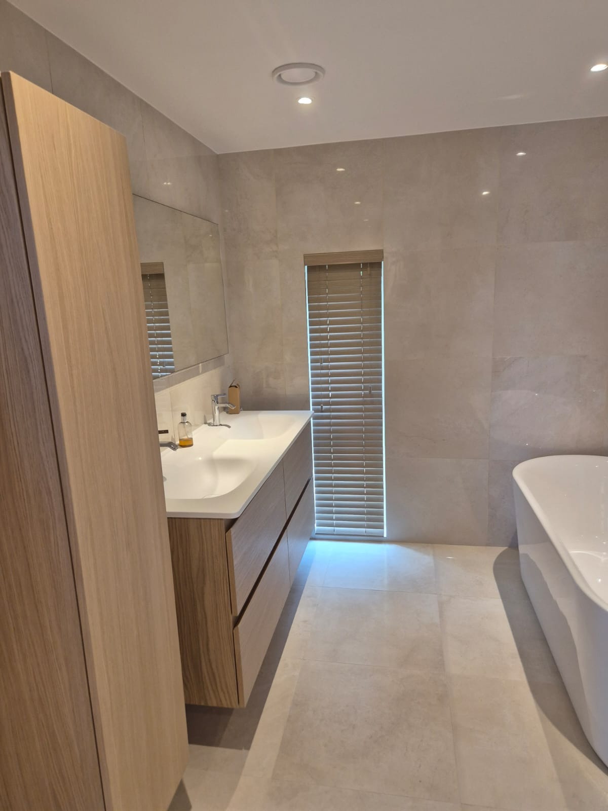 Jasna łazienka z drewnianą szafką pod umywalką, beżowymi płytkami i białą wanną. Okno z żaluzjami w tle. Minimalistyczny styl.