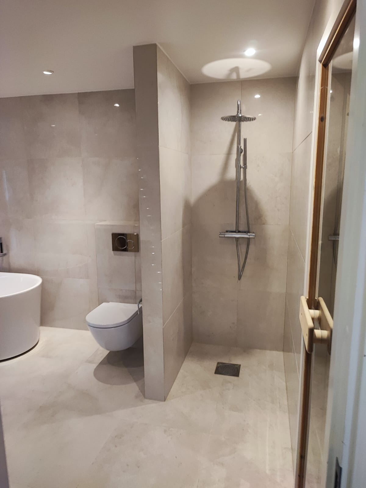 Jasna łazienka z kabiną prysznicową typu walk-in, toaletą podwieszaną i fragmentem wanny. Minimalistyczny design, beżowe płytki, punktowe oświetlenie.