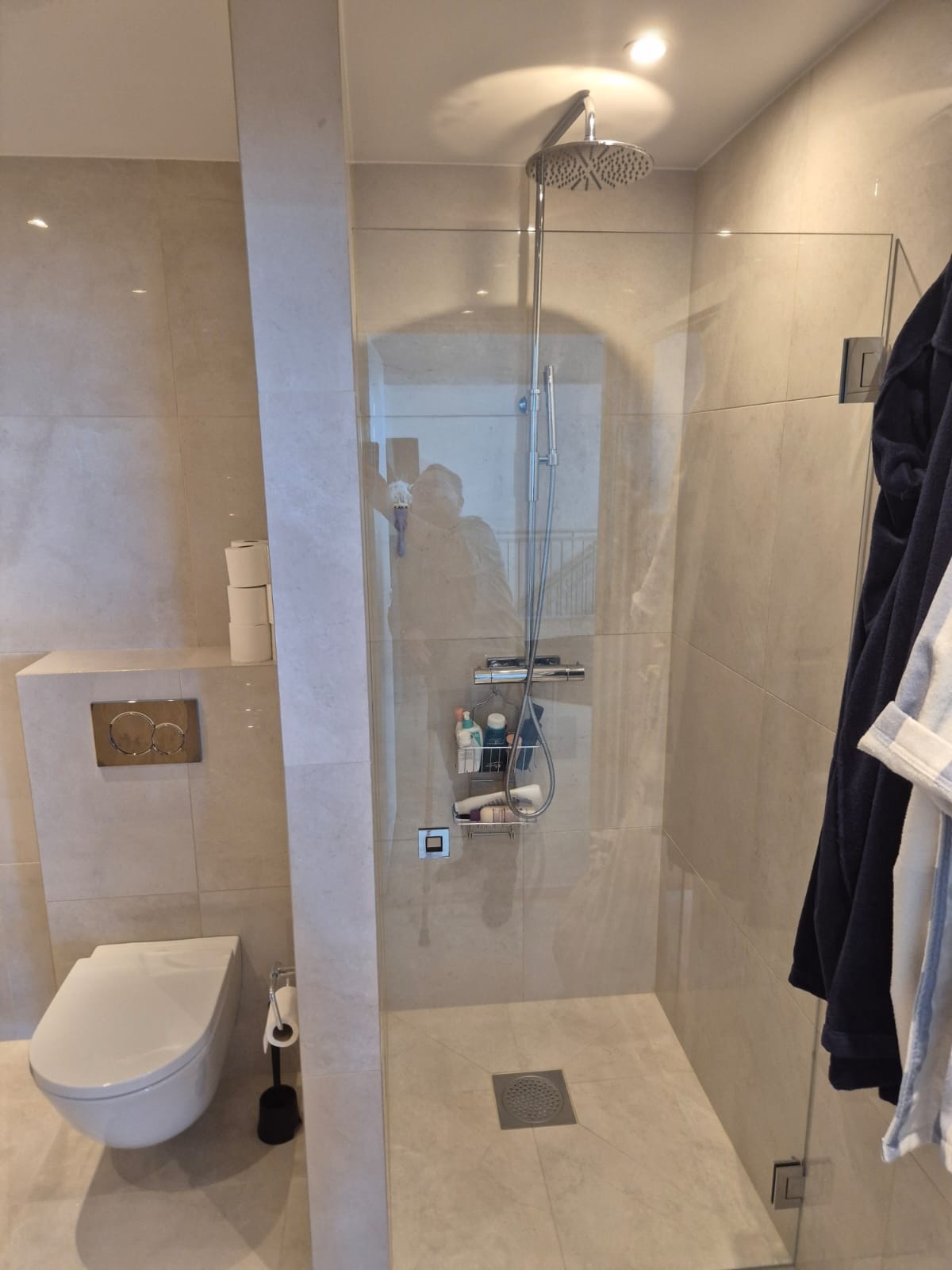 Nowoczesna łazienka z kabiną prysznicową typu walk-in, beżowe płytki, podwieszana toaleta i chromowana armatura. Minimalistyczny design z akcentami na detale.