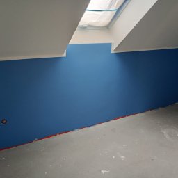 mitrahome serwis spółka cywilna Dorota Garczyńska Agnieszka Knapek - Pomieszczenie po malowaniu na niebiesko, widoczne zabezpieczone okno dachowe i szary betonowy podkład. Czerwona taśma zabezpiecza dolną krawędź ściany.