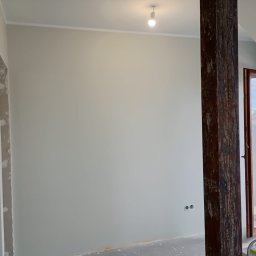 mitrahome serwis spółka cywilna Dorota Garczyńska Agnieszka Knapek - Wnętrze po malowaniu ścian na jasny odcień szarości, widoczna drewniana belka i okno balkonowe z widokiem na zewnątrz. Świeżo pomalowane gniazdka elektryczne.