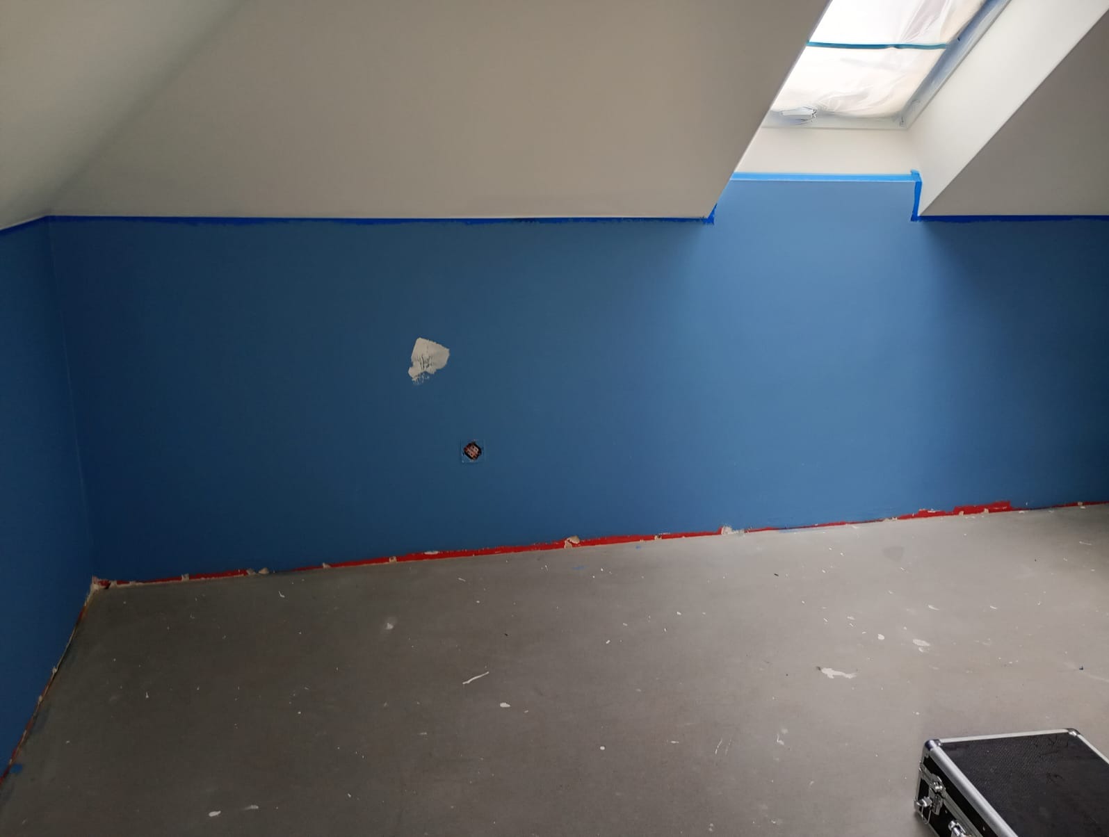 Ściana w kolorze niebieskim, zabezpieczona taśmą malarską przy suficie i podłodze, widoczne ślady po szpachlowaniu i gniazdko elektryczne. Podłoga betonowa.