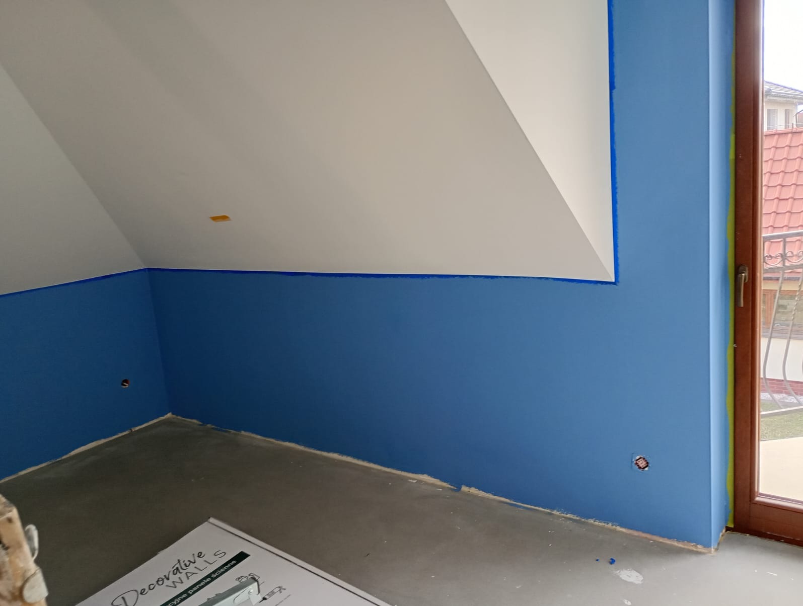 Pomalowane na niebiesko poddasze z białym sufitem, zabezpieczone taśmą malarską przy oknie. Częściowo widoczne panele dekoracyjne. Widok na zewnątrz przez okno.