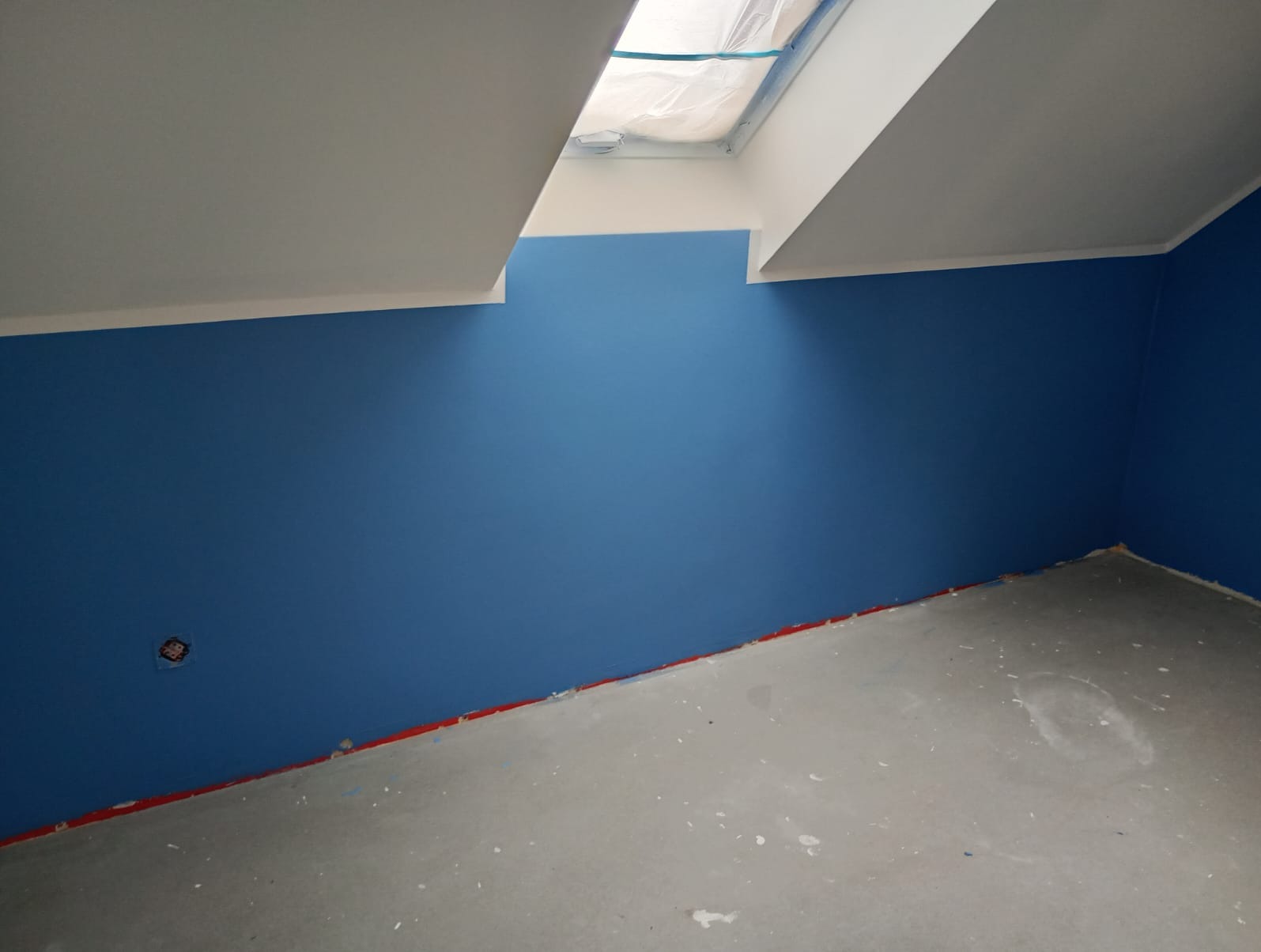 Pomieszczenie po malowaniu na niebiesko, widoczne zabezpieczone okno dachowe i szary betonowy podkład. Czerwona taśma zabezpiecza dolną krawędź ściany.