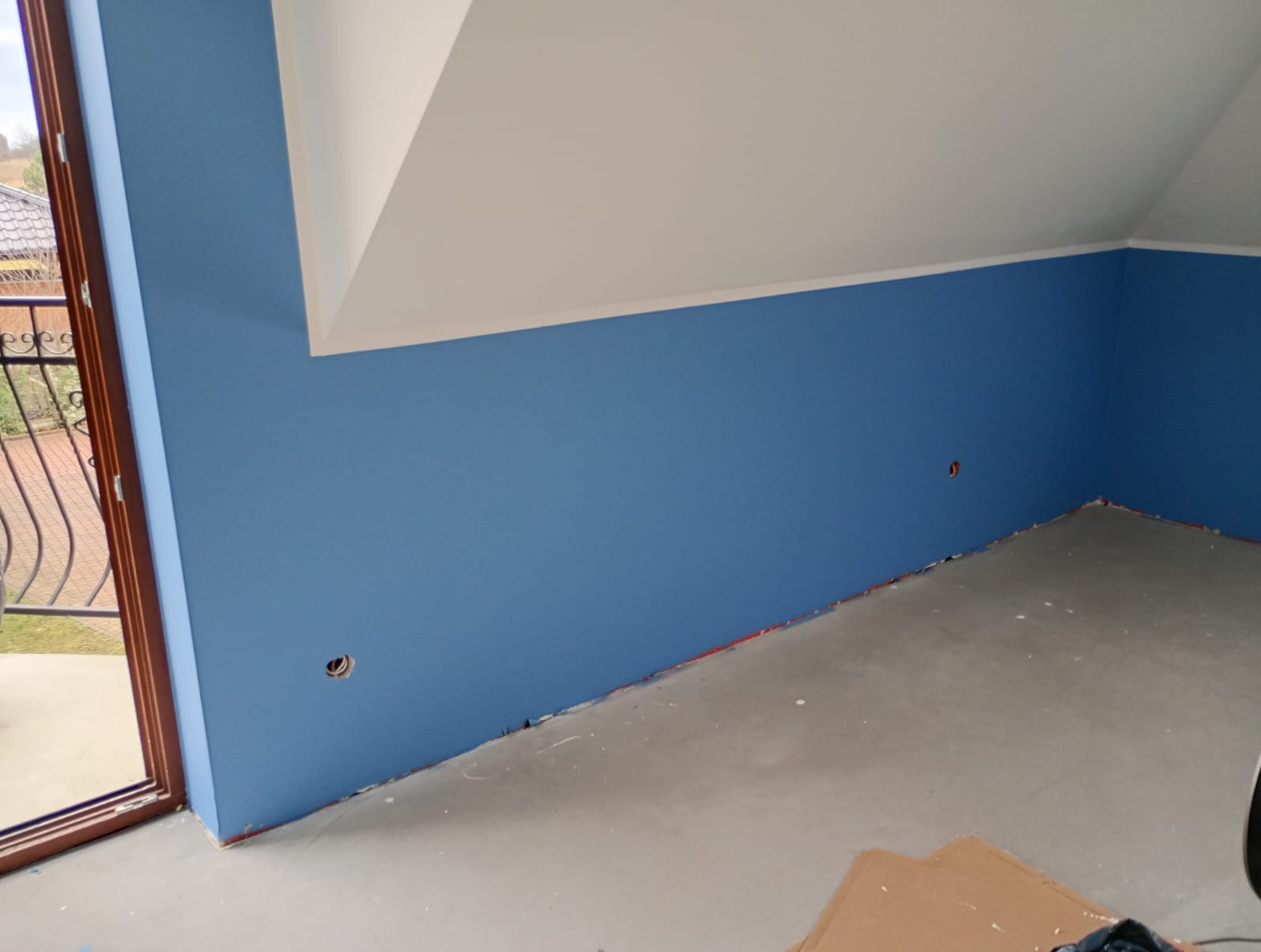 Pomieszczenie po malowaniu ścian na niebiesko, widoczne gładkie powierzchnie i przygotowane otwory na gniazdka, sufit biały, okno z widokiem na zewnątrz.