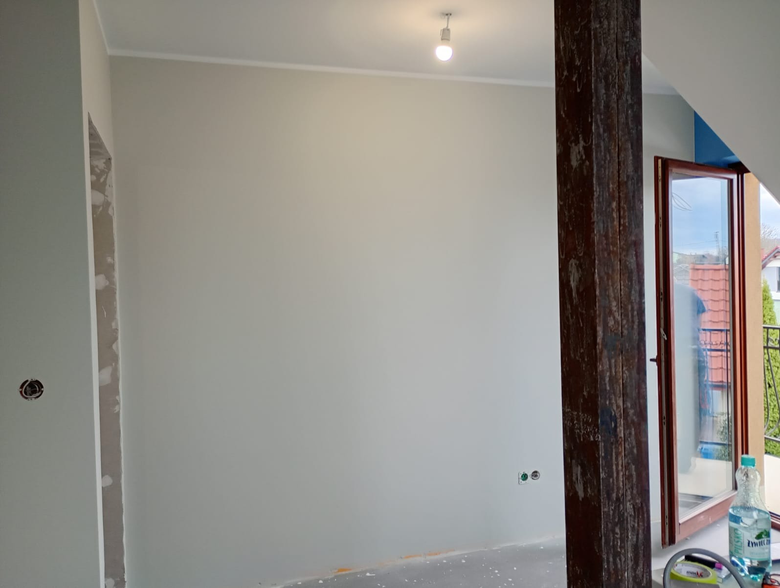 Wnętrze po malowaniu ścian na jasny odcień szarości, widoczna drewniana belka i okno balkonowe z widokiem na zewnątrz. Świeżo pomalowane gniazdka elektryczne.