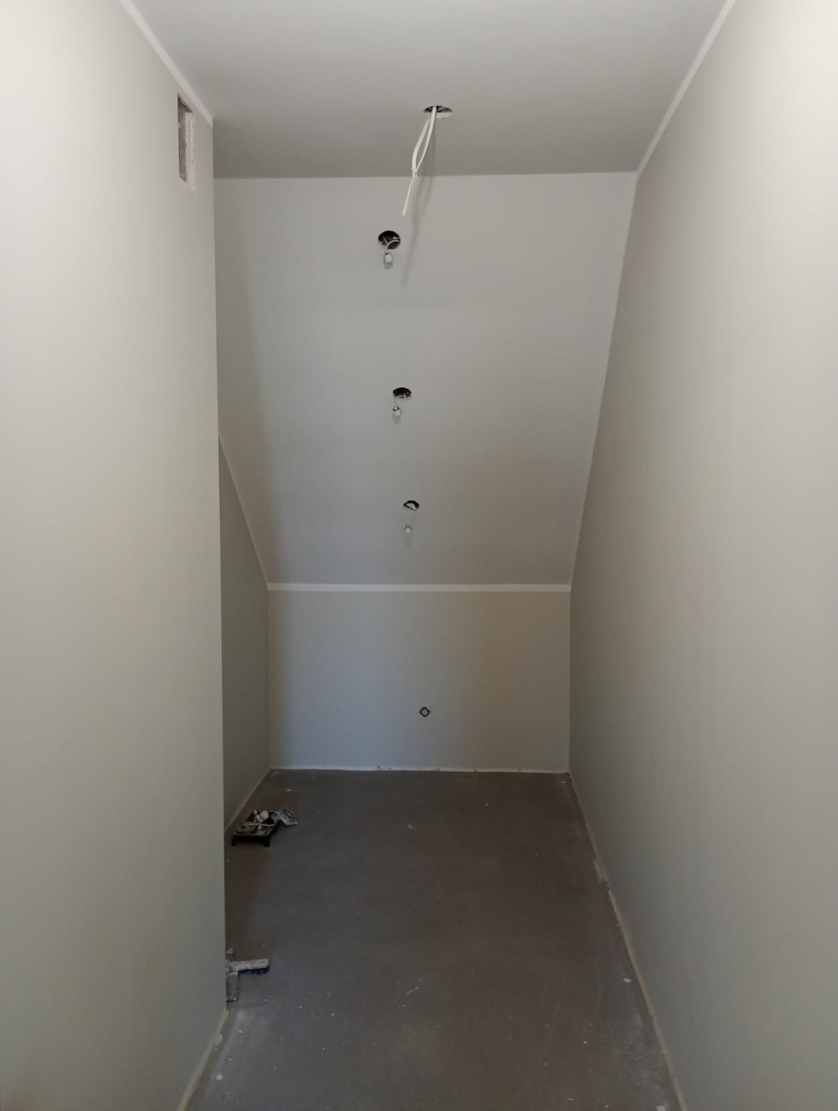 Wnętrze poddasza po szpachlowaniu i malowaniu na neutralny odcień szarości, z widocznymi otworami na oświetlenie i narzędziami malarskimi w rogu.