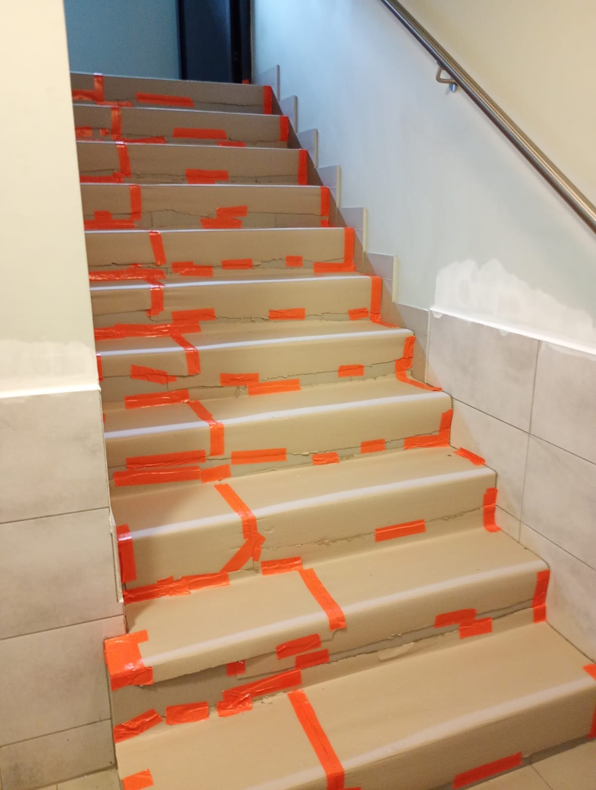 Schody zabezpieczone pomarańczową taśmą malarską i papierem przed malowaniem, widoczne płytki na ścianie i ślady po szpachlowaniu. Przygotowanie do prac wykończeniowych.