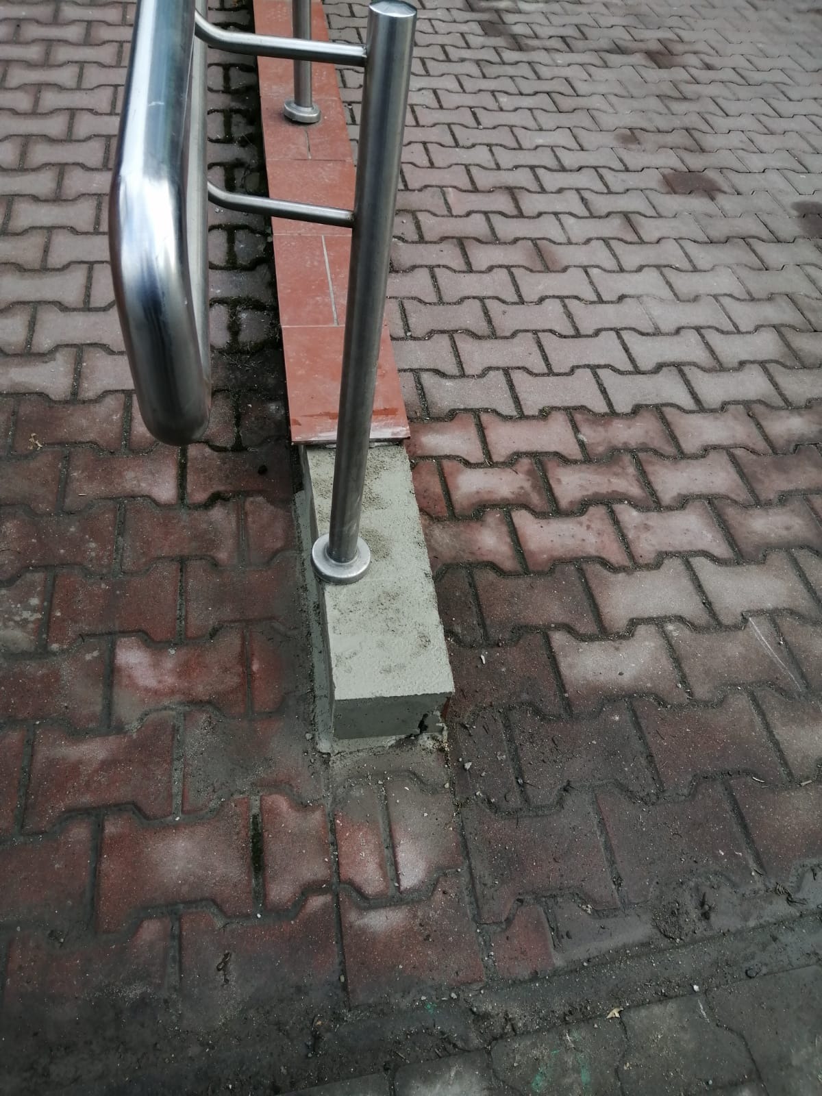 Naprawiony fragment murku przy metalowej barierze na chodniku z kostki brukowej. Widoczne świeże zaprawy i fugi. Prace remontowe na zewnątrz.