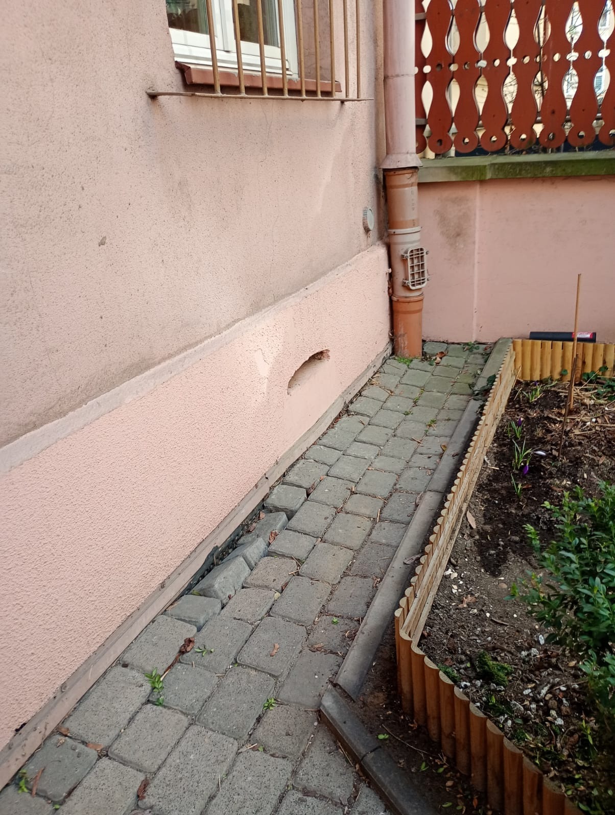Fragment elewacji budynku z różową strukturą, obok chodnik z kostki brukowej i rabata z drewnianym obrzeżem. Widoczna rynna i okno z kratą.