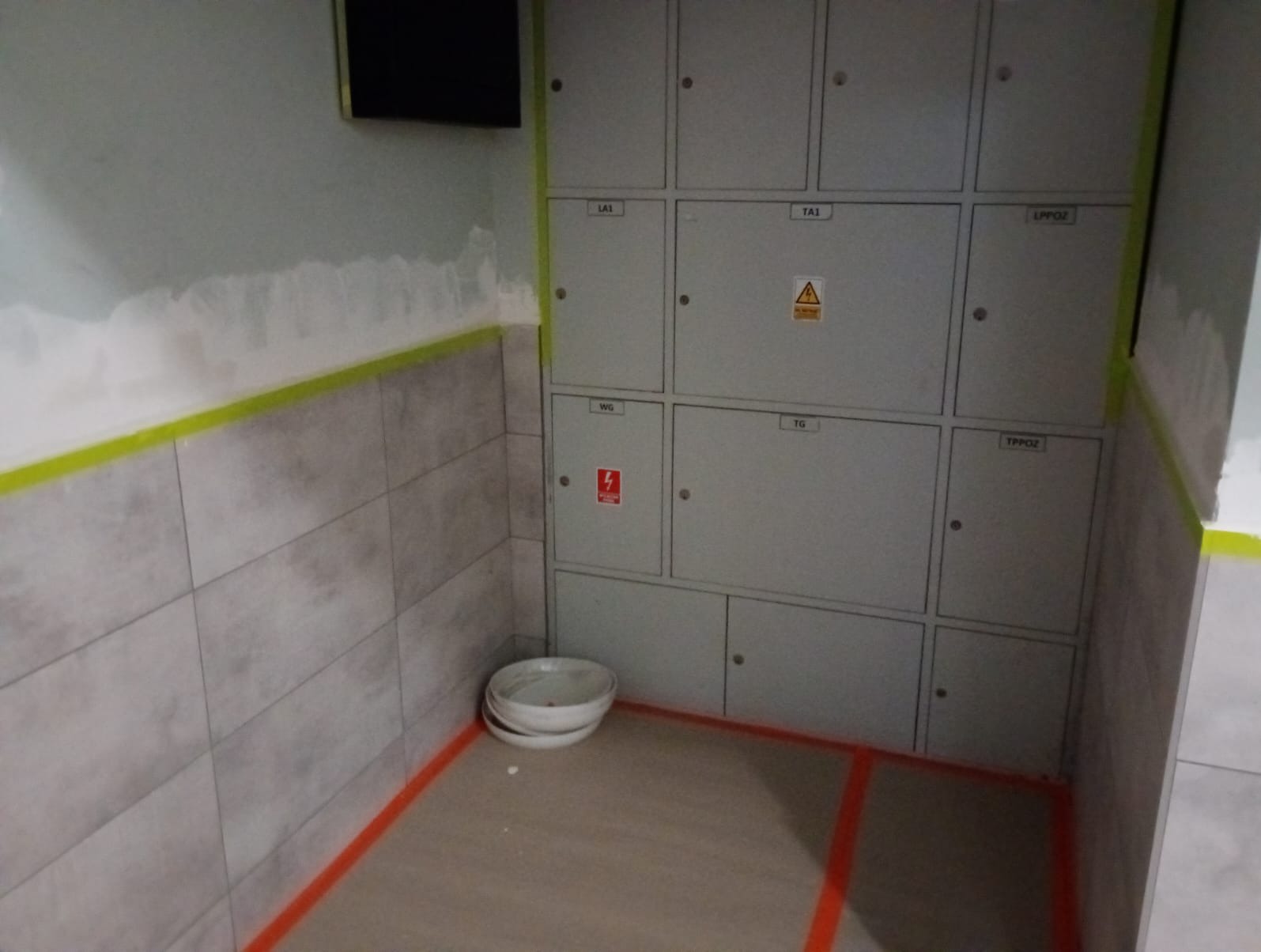 Ściana z szpachlowanymi ubytkami, zabezpieczona taśmą malarską, obok szare płytki i białe miski. Na ścianie szafki elektryczne.