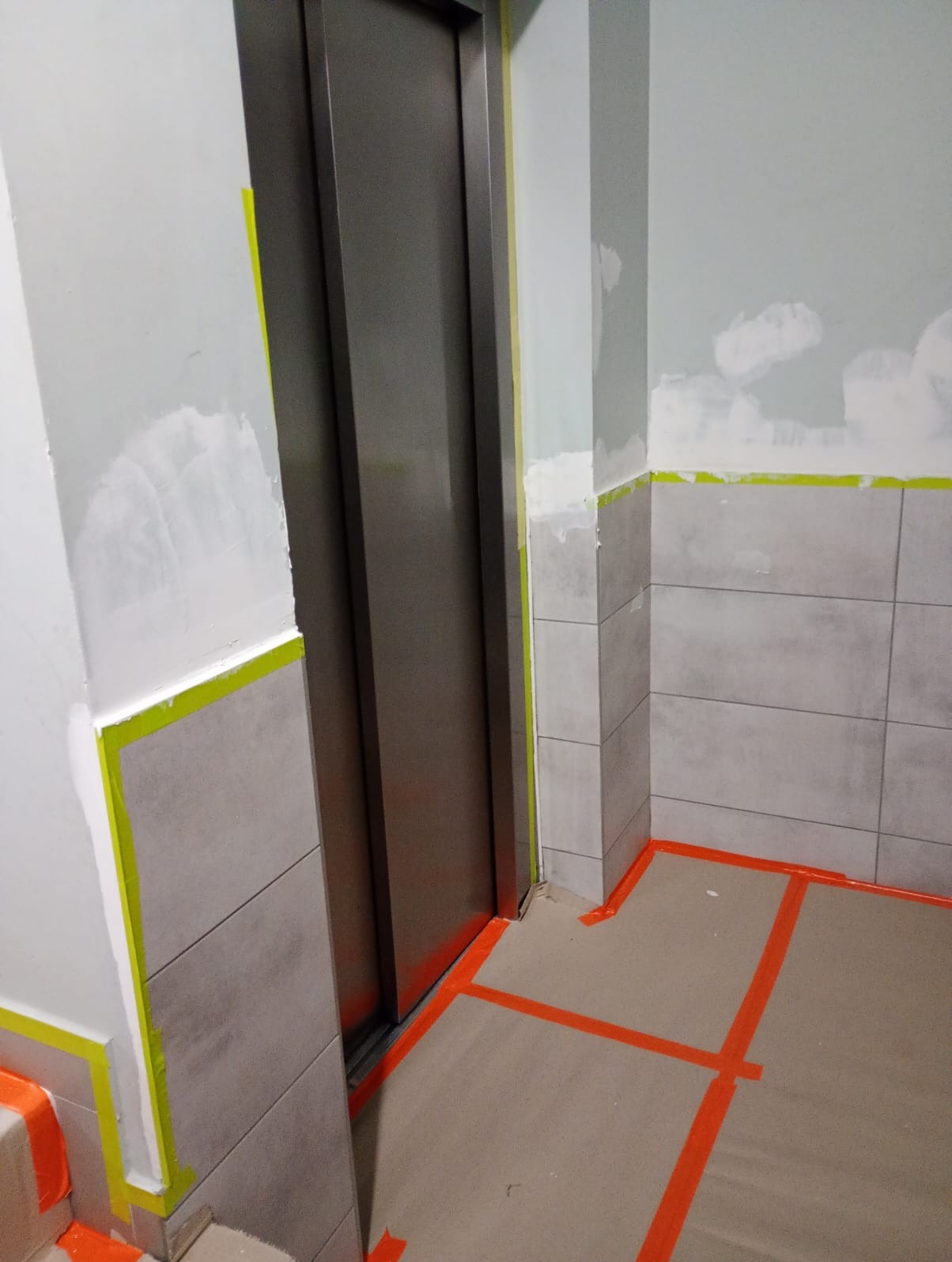 Korytarz przed windą w trakcie prac wykończeniowych: ściany z fragmentami szpachli, zabezpieczone taśmą podłogi i płytki. Widoczny etap przygotowania do malowania.