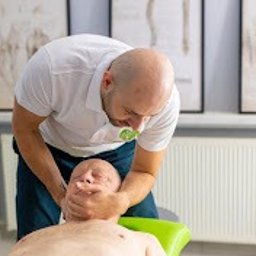 Synergia Centrum Żary - Fizjoterapia - Rehabilitacja - Masaż - Fizjoterapeuta delikatnie masuje i rozluźnia mięśnie szyi starszego mężczyzny leżącego na zielonym leżaku, w tle biały grzejnik i grafiki na ścianie.