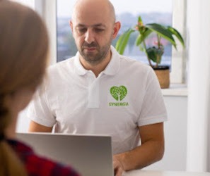 Mężczyzna w białej koszulce polo z logo firmy Synergia, pochylony nad laptopem, konsultuje się z pacjentem siedzącym naprzeciwko, widok zza pleców pacjenta.