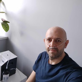 Mężczyzna z kaniulą nosową, podłączony do koncentratora tlenu stojącego na stoliku przy ścianie, obok doniczka z rośliną.