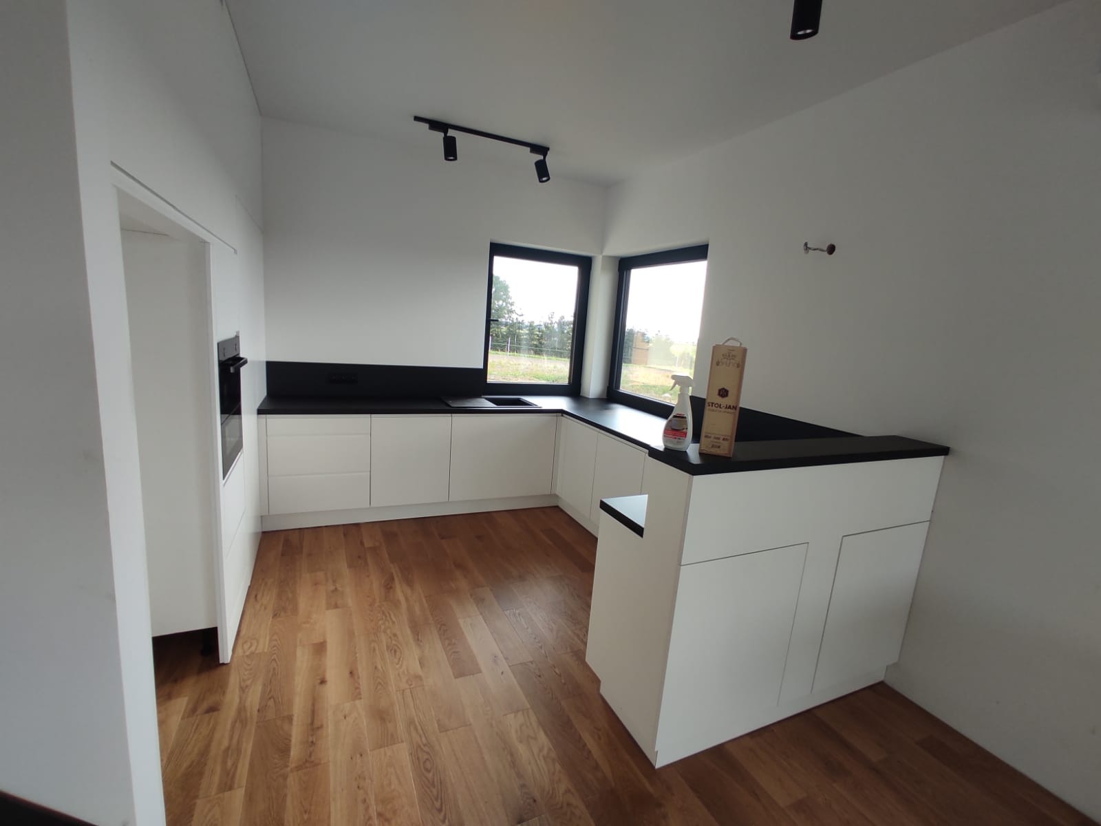 Nowoczesna kuchnia z białymi szafkami i czarnym blatem, widok na zewnątrz przez narożne okno, drewniana podłoga, minimalistyczne oświetlenie sufitowe.