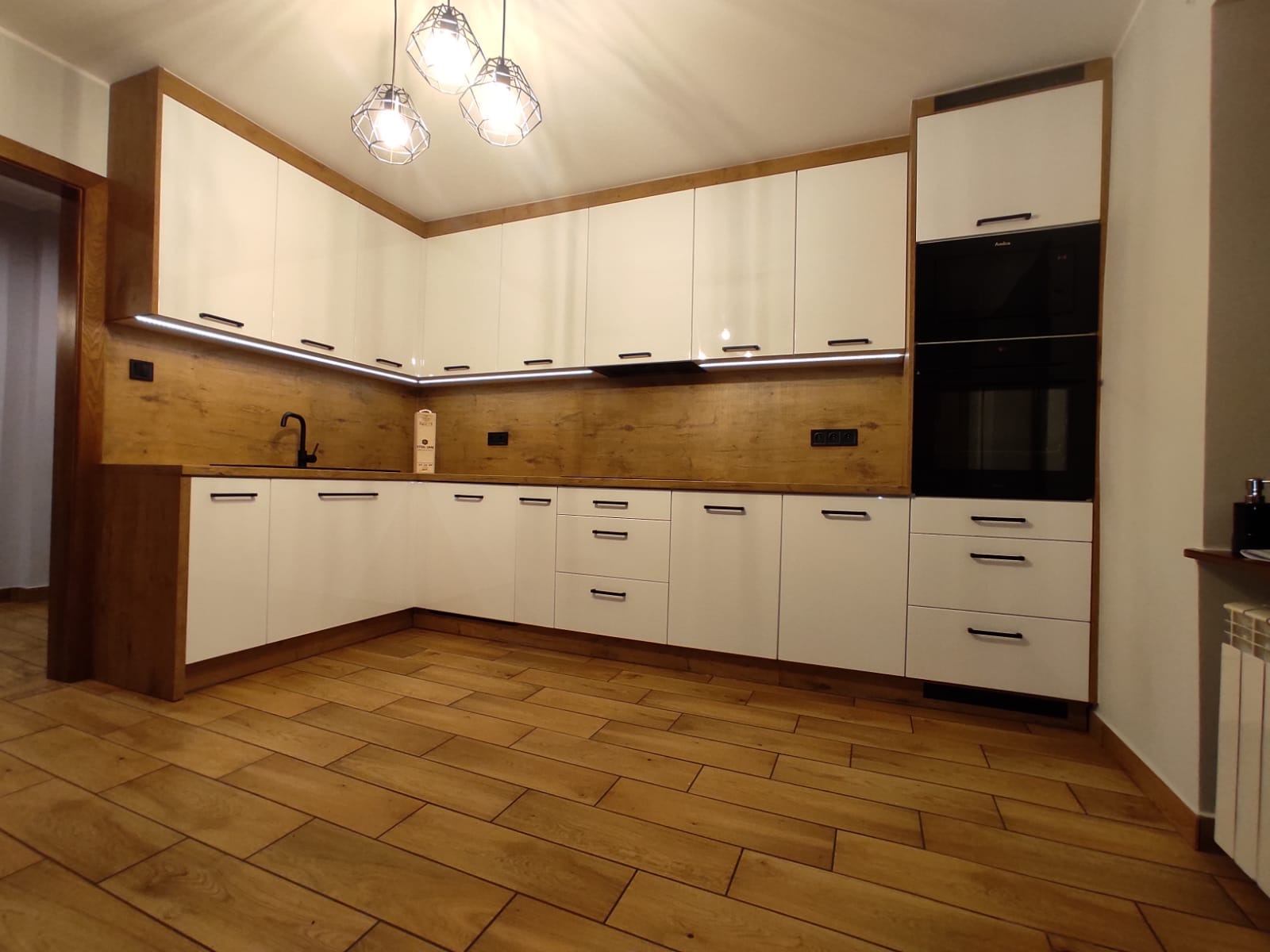 Nowoczesna kuchnia z białymi, lakierowanymi frontami, drewnianym blatem i podświetleniem LED, uzupełniona o czarne akcenty w postaci baterii i uchwytów.