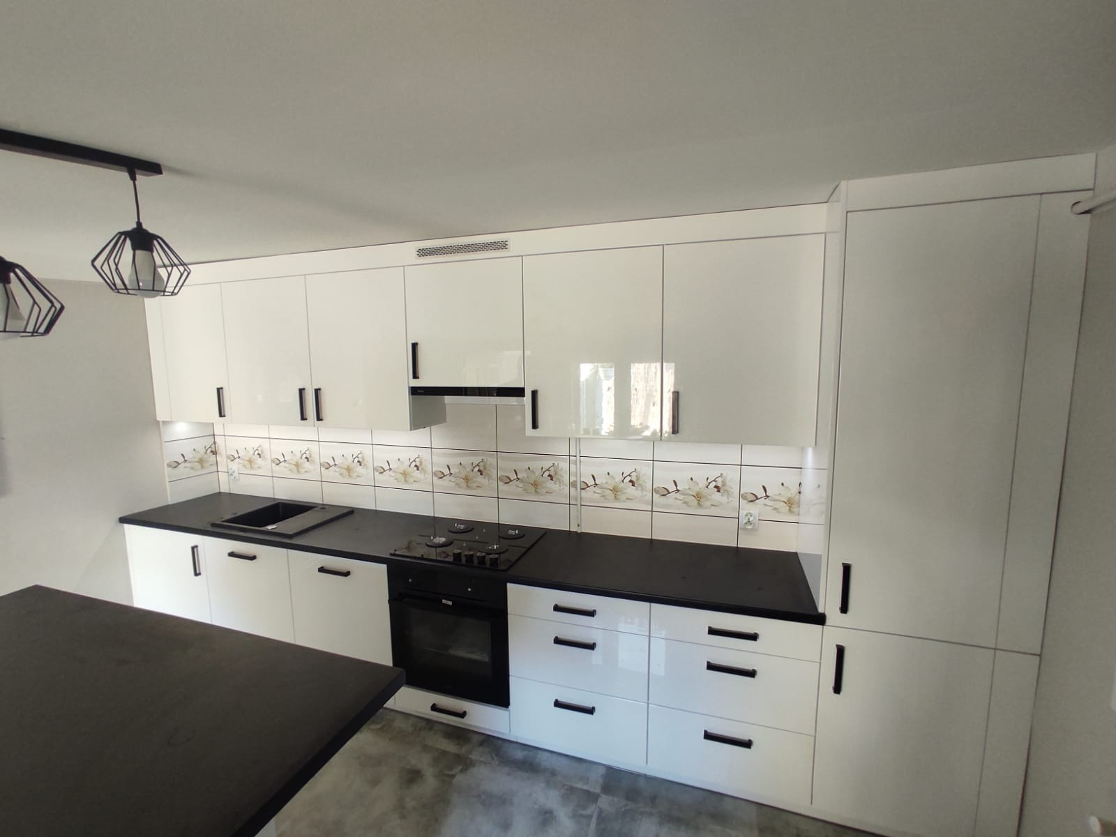 Biała kuchnia z czarnym blatem i frontami bez uchwytów, płytki dekoracyjne nad blatem, nowoczesne lampy wiszące.