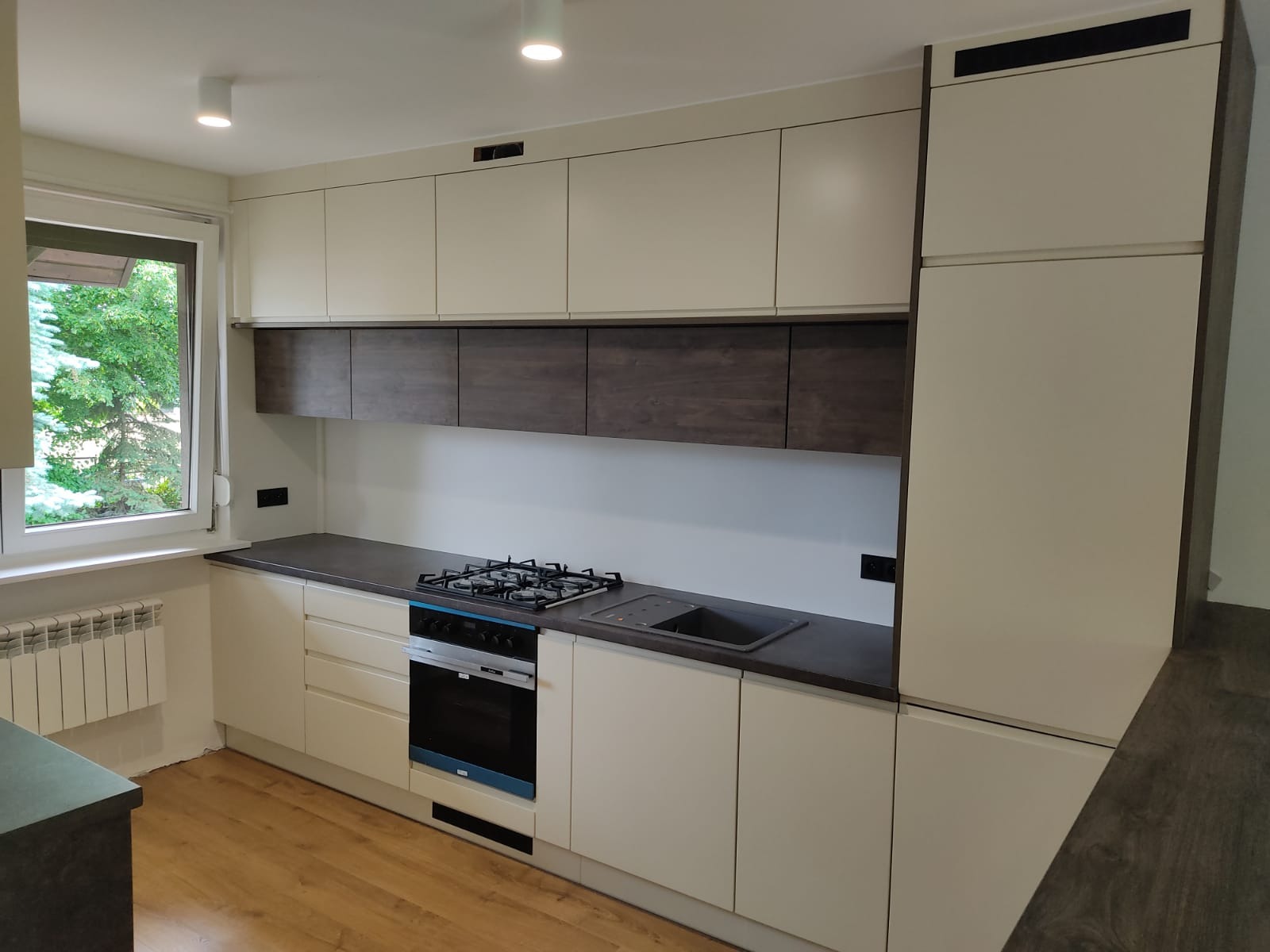 Jasna kuchnia z górnymi szafkami w kolorze ecru i dolnymi w odcieniu ciemnego drewna, z widoczną płytą gazową i piekarnikiem, oświetlona światłem dziennym wpadającym przez okno.